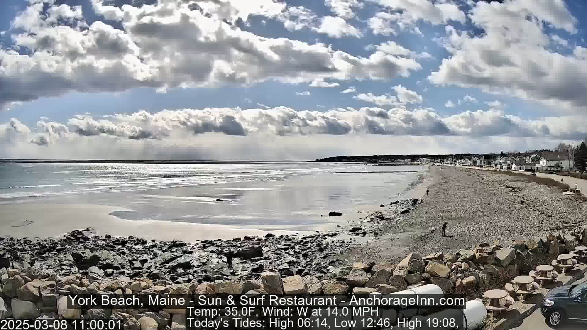 A partly cloudy day reveals a wide sandy beach with rocky areas, a calm ocean, and a shoreline of houses visible in the distance.