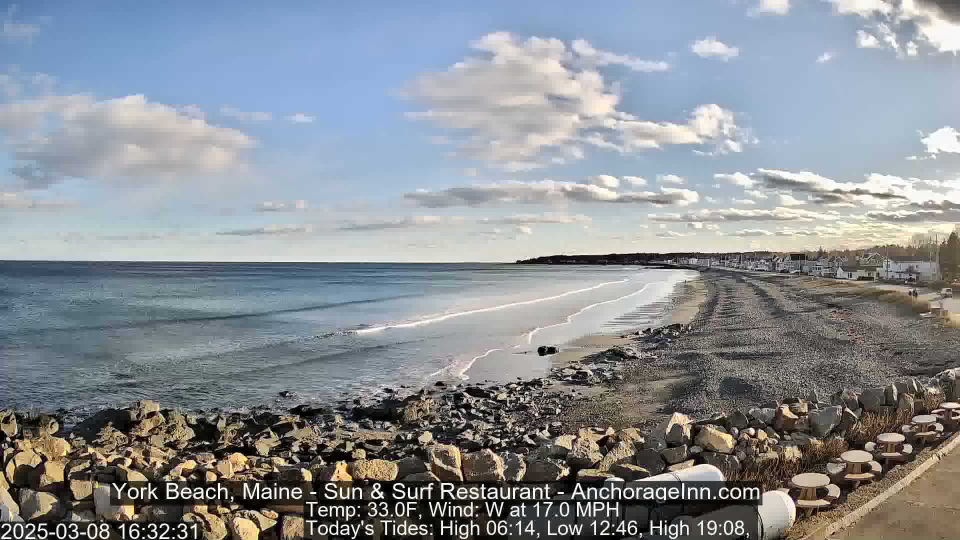 A long, sandy beach stretches from a rocky shoreline toward a small town under a partly cloudy sky, with gentle waves lapping the shore.