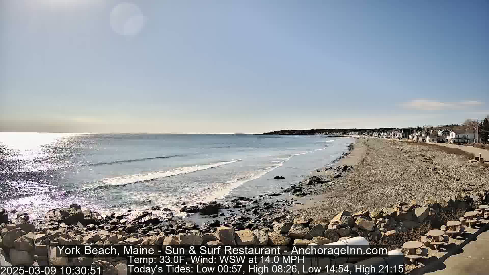 A sandy beach with a rocky shoreline stretches along a calm ocean under a sunny sky.