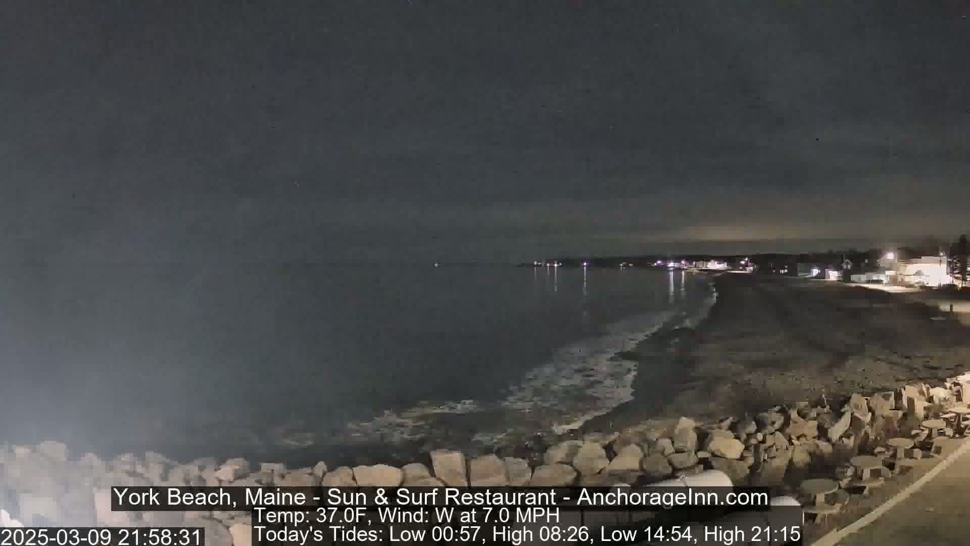 A dark, nighttime view of a rocky shoreline and calm ocean under a mostly clear sky.