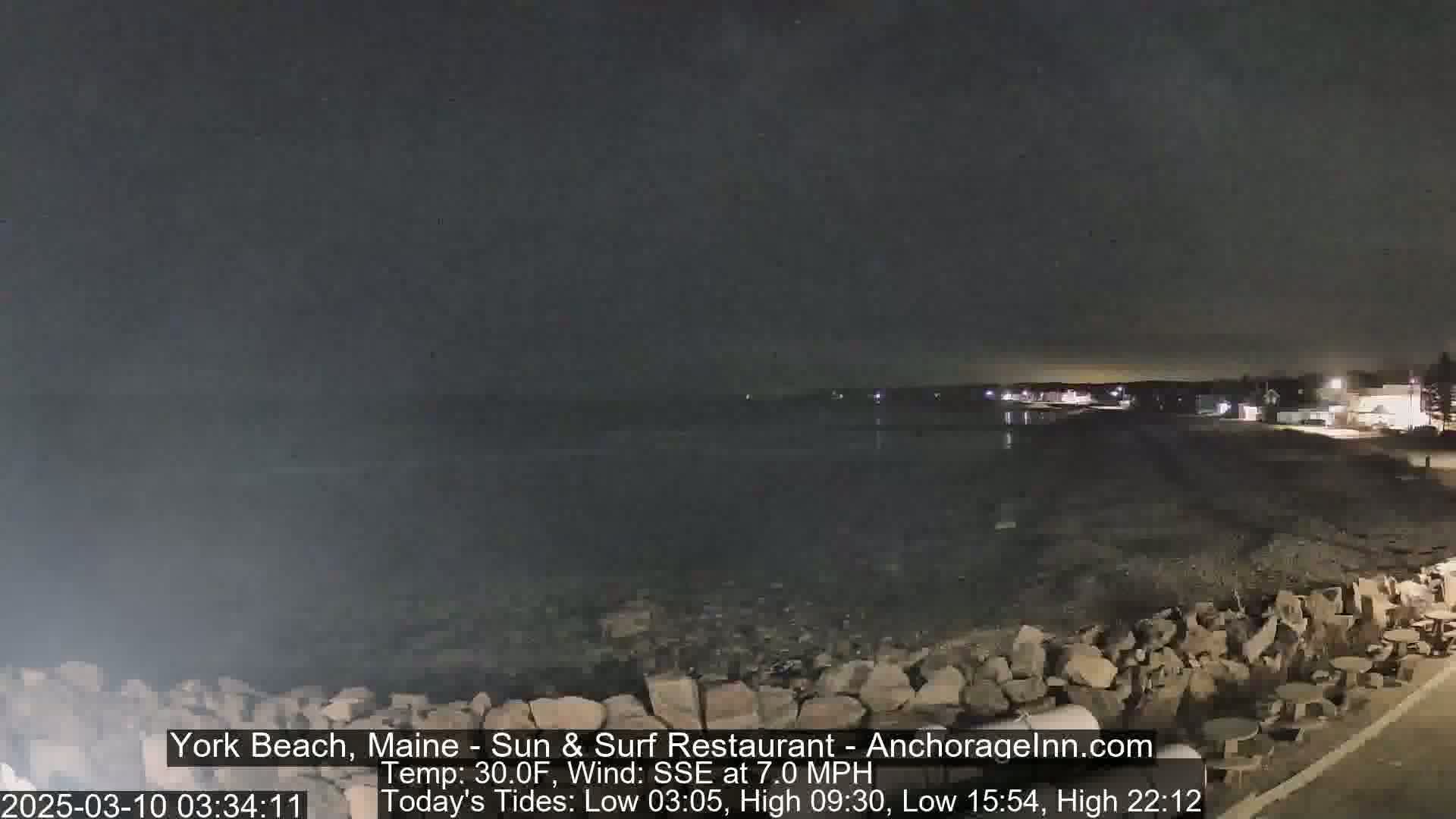 A dark nighttime scene shows a rocky shoreline and calm ocean water under a clear sky.