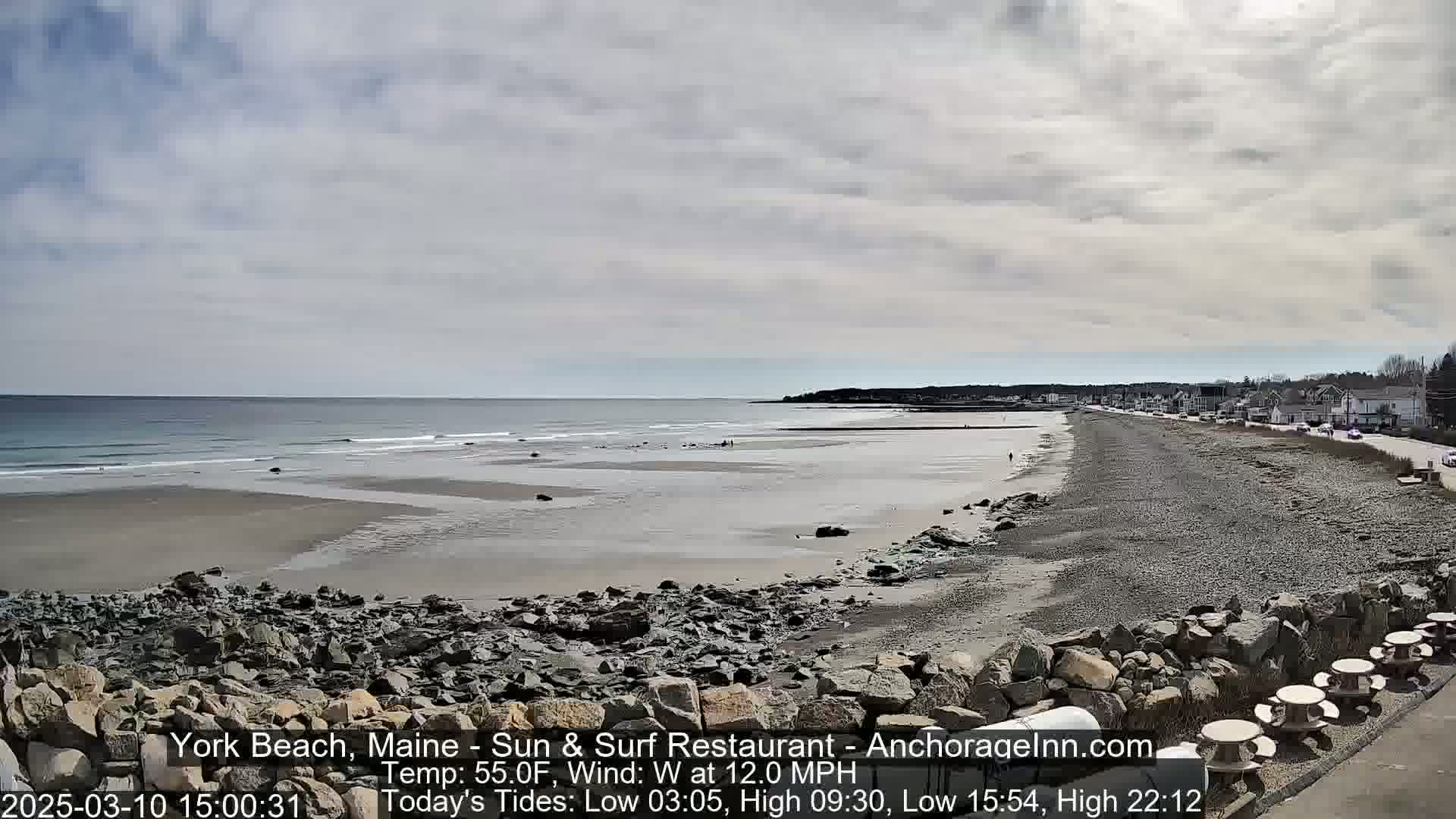 A wide sandy beach stretches along a rocky coastline under a partly cloudy sky, with gentle waves lapping the shore and a road visible beyond.