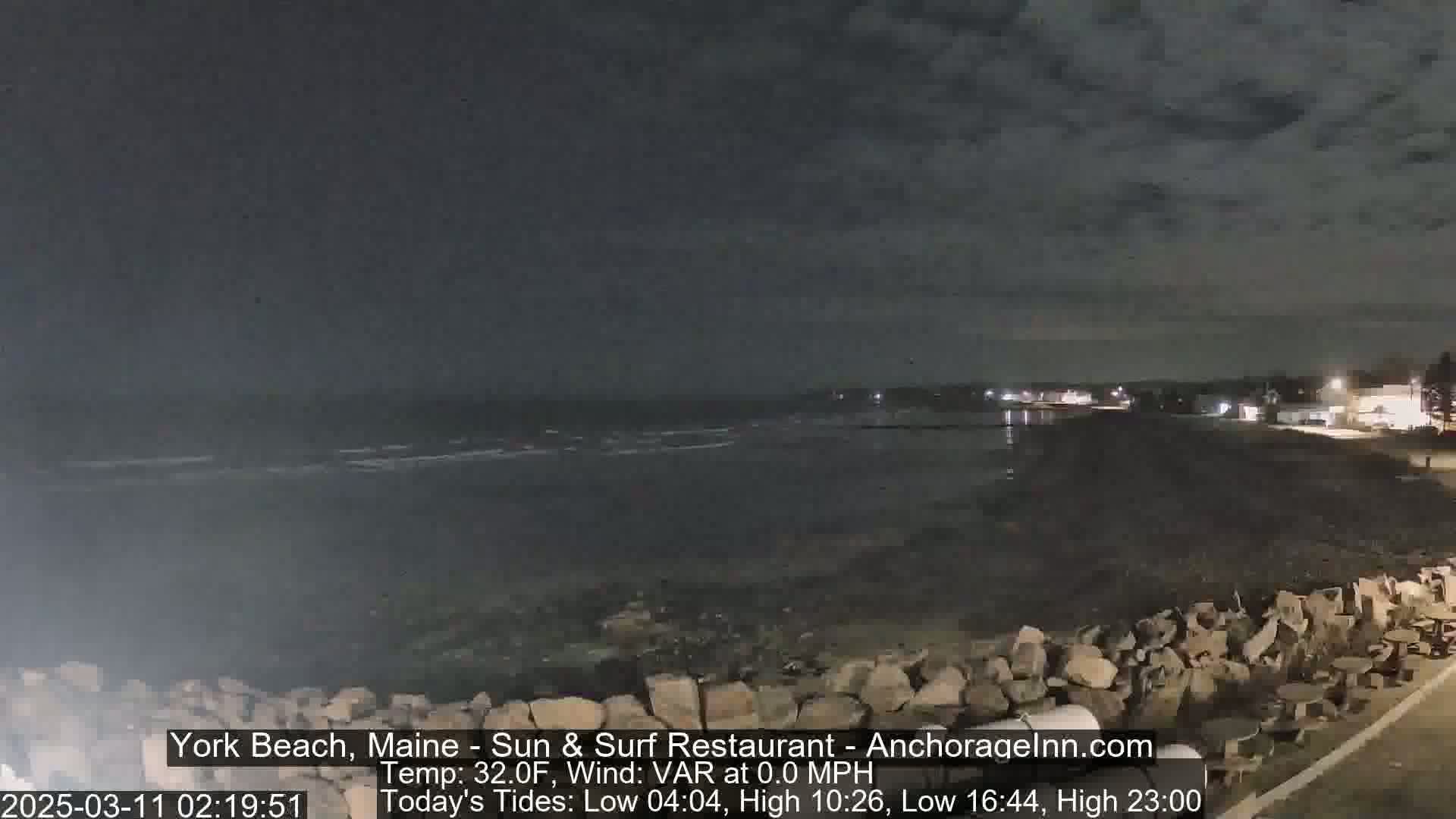 A nighttime view of a rocky shoreline with calm water under a partly cloudy sky.