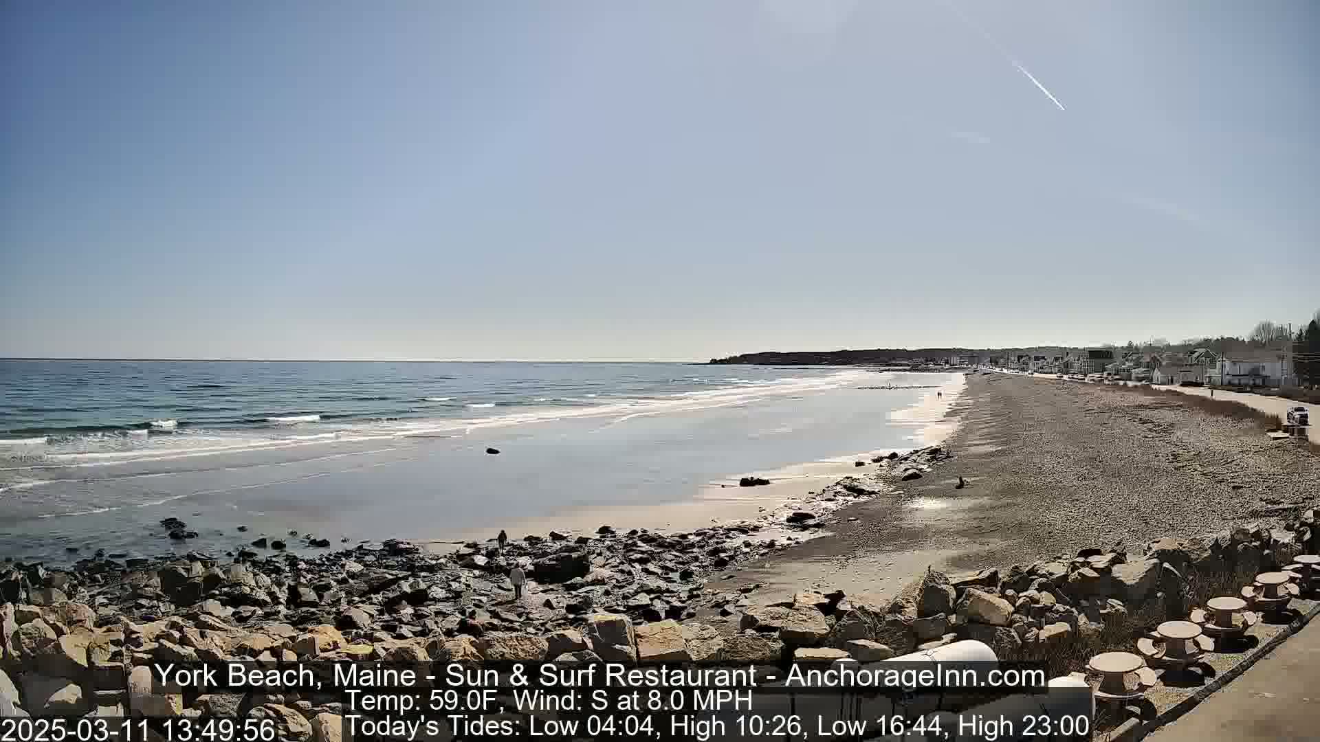 A sandy beach with rocky outcrops and calm waves under a clear blue sky stretches to a shoreline lined with buildings.