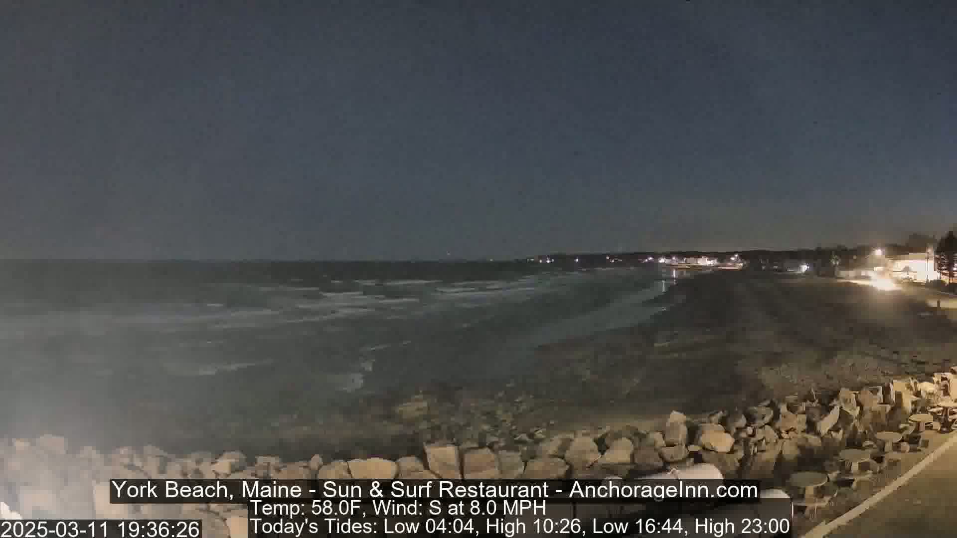 A dark, nighttime beach scene shows a rocky shoreline and calm ocean waves under a clear, dark sky.