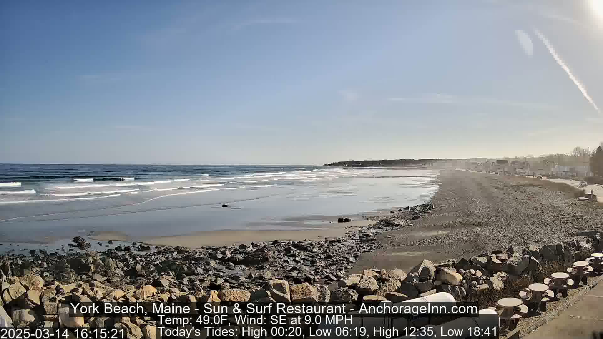 A rocky beach stretches alongside a calm ocean under a clear blue sky, with small waves breaking gently on the shore.