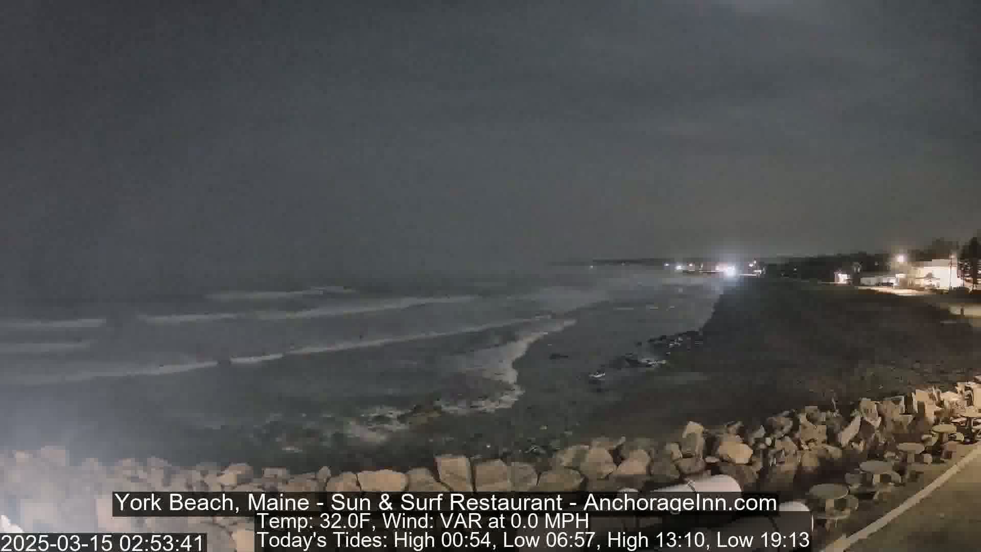 A dark, nighttime beach scene shows a calm ocean with small waves lapping the shore near a rocky embankment; a few lights are visible in the distance.