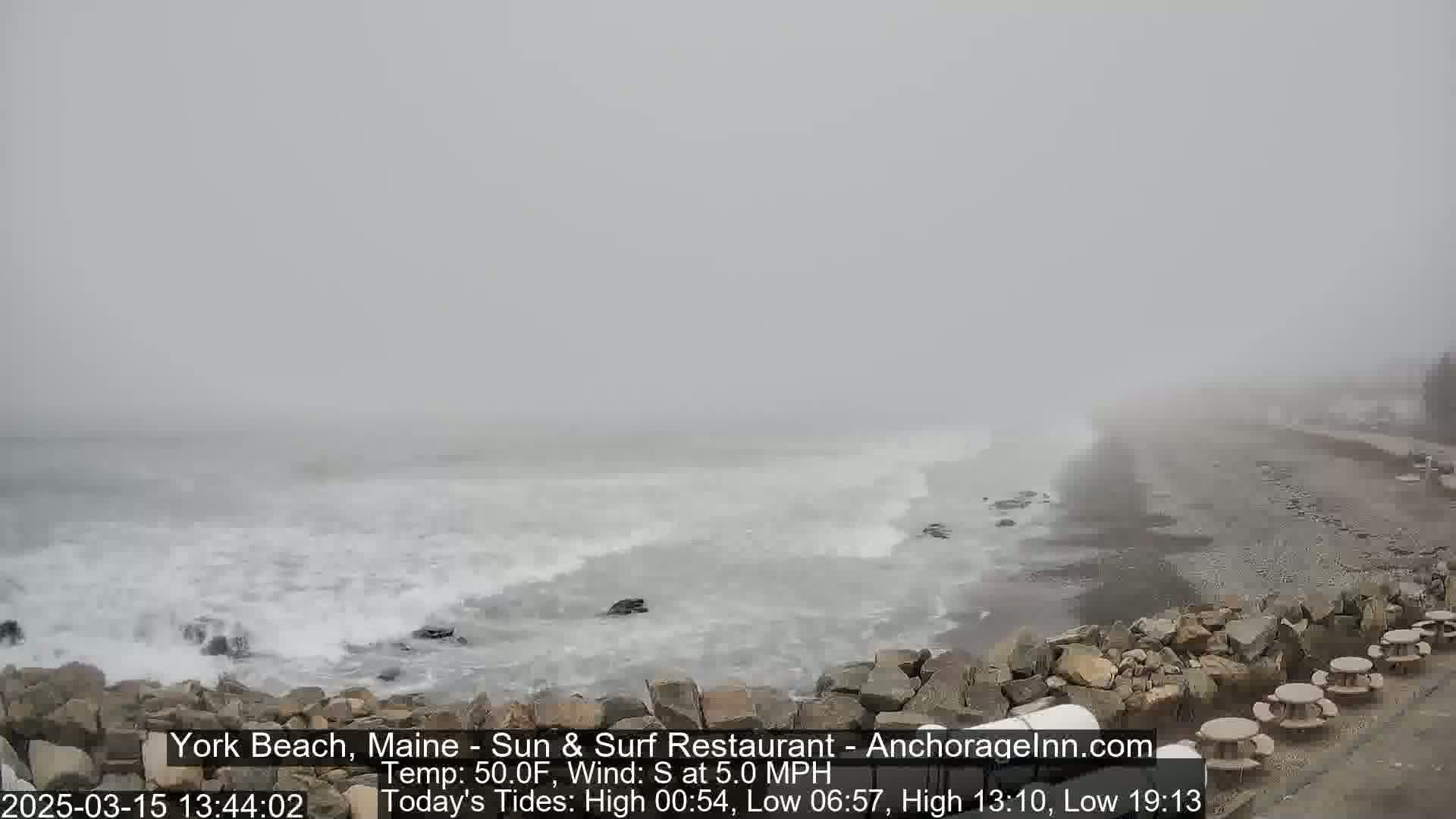 A foggy beach scene shows waves crashing on a rocky shore near picnic tables.