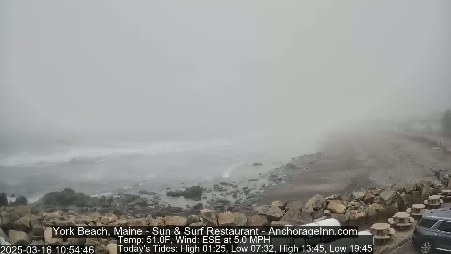 A foggy beach scene shows a rocky shoreline meeting a gray, wavy ocean under a thick fog.