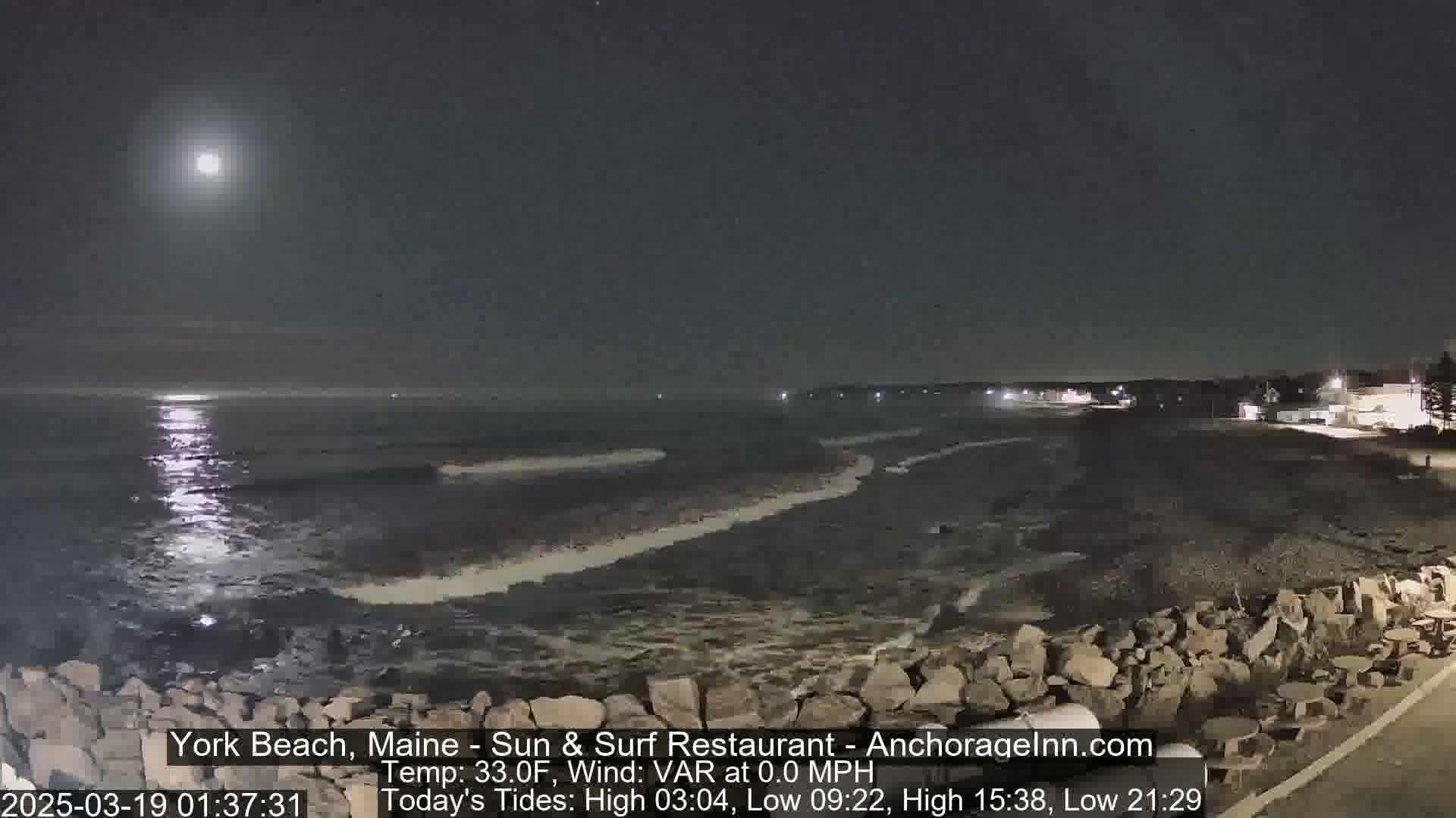 A nighttime view of a rocky shoreline and ocean under a bright moon, with calm water.