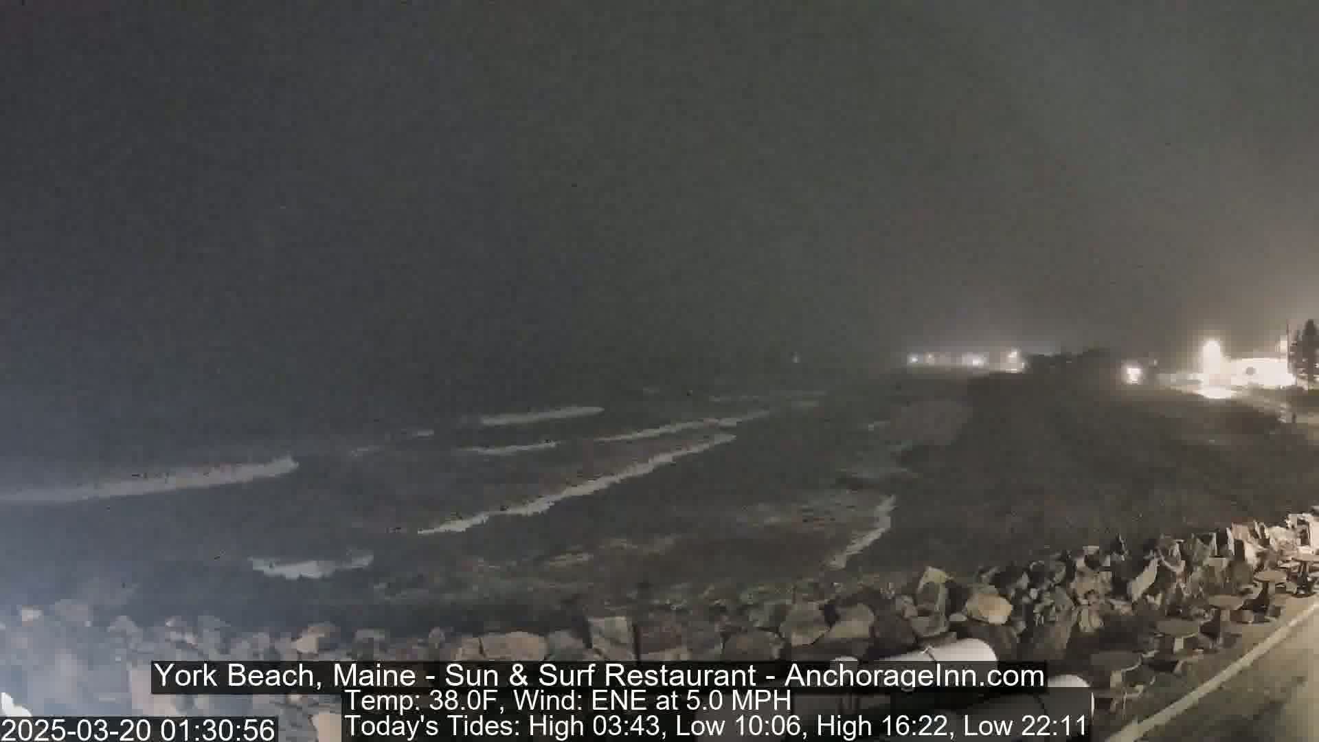 A dark, nighttime beach scene shows a rocky shoreline with waves lapping against it under a slightly overcast sky, with a faint wind.