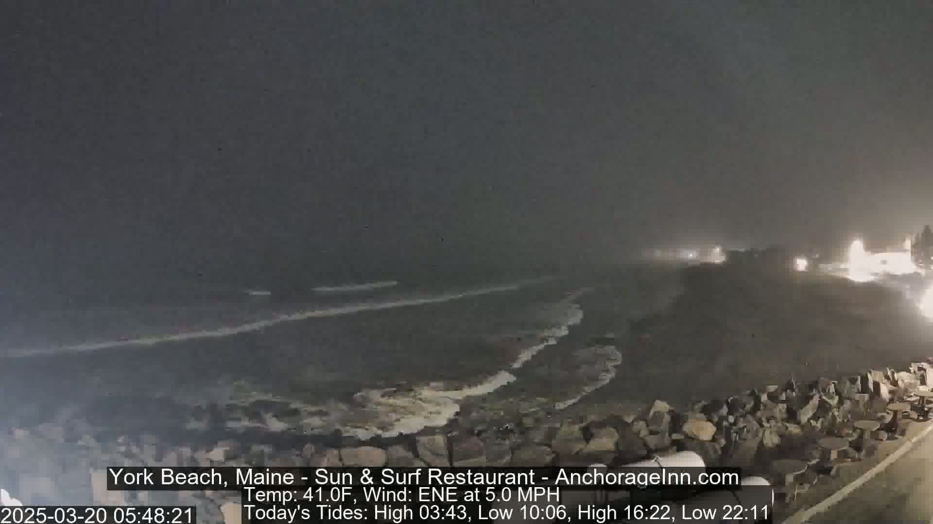 A dark, nighttime view of a rocky coastline with waves crashing on the shore under a hazy sky.