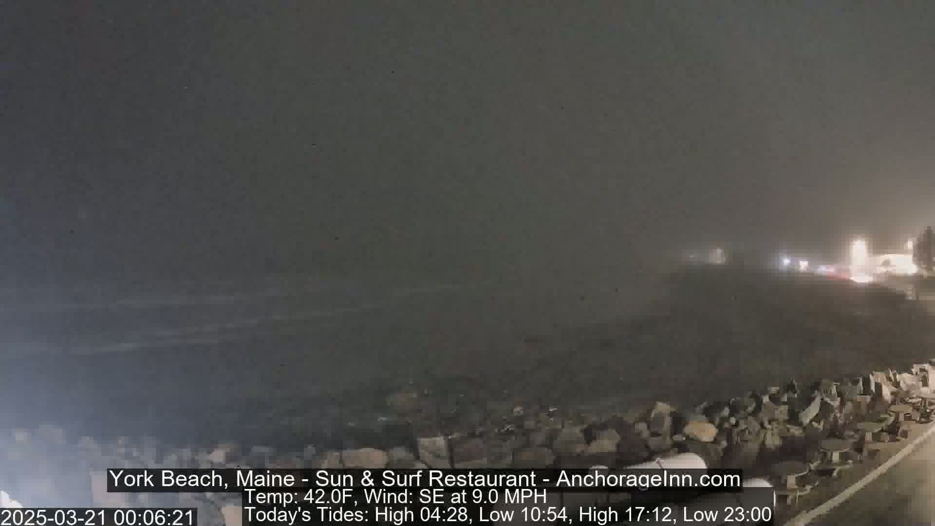 A foggy, nighttime coastal scene shows a rocky shoreline with a faint view of lights in the distance.