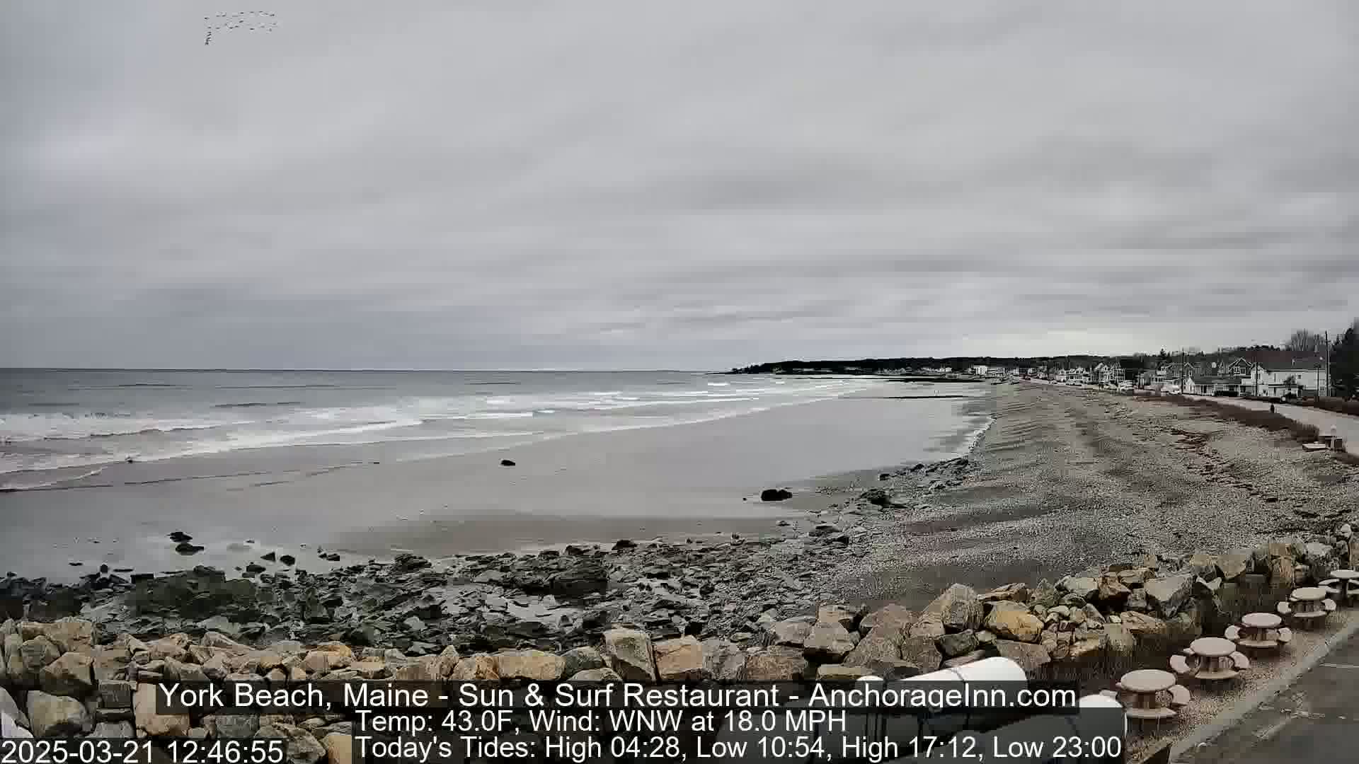 A wide, sandy beach with rocky shoreline stretches along a calm ocean under an overcast sky, with a road and buildings visible in the distance.