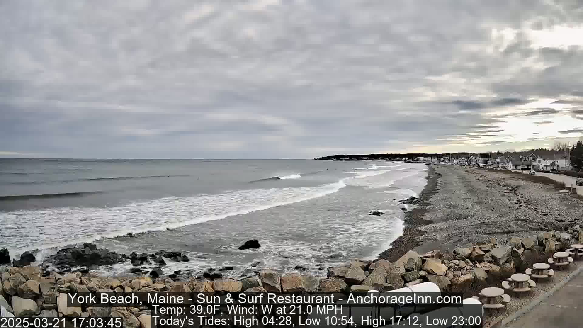 A beach with gray, overcast skies and a 21 mph westerly wind shows gentle waves lapping a rocky shore, with buildings visible in the distance.