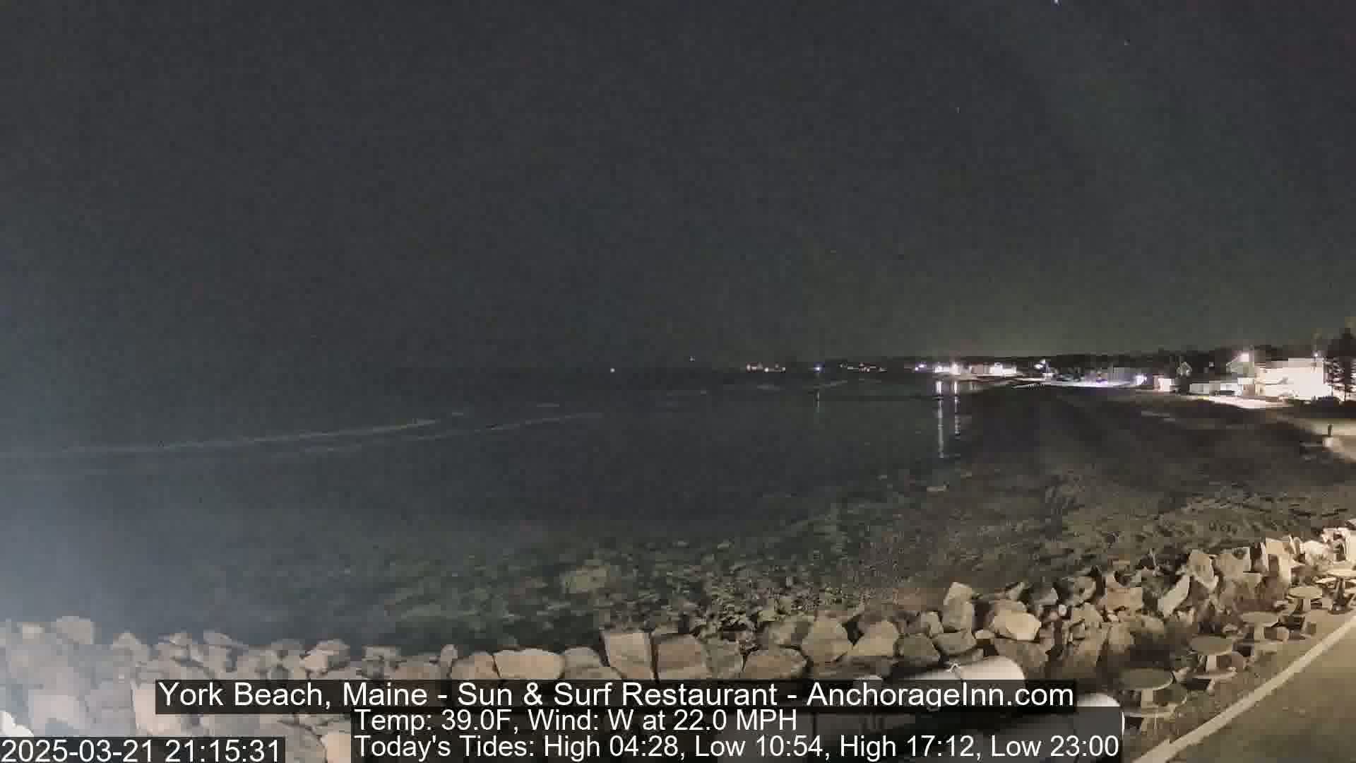 A dark, nighttime beach scene with a rocky shoreline and calm water under a clear, starlit sky, with a moderate westerly wind.