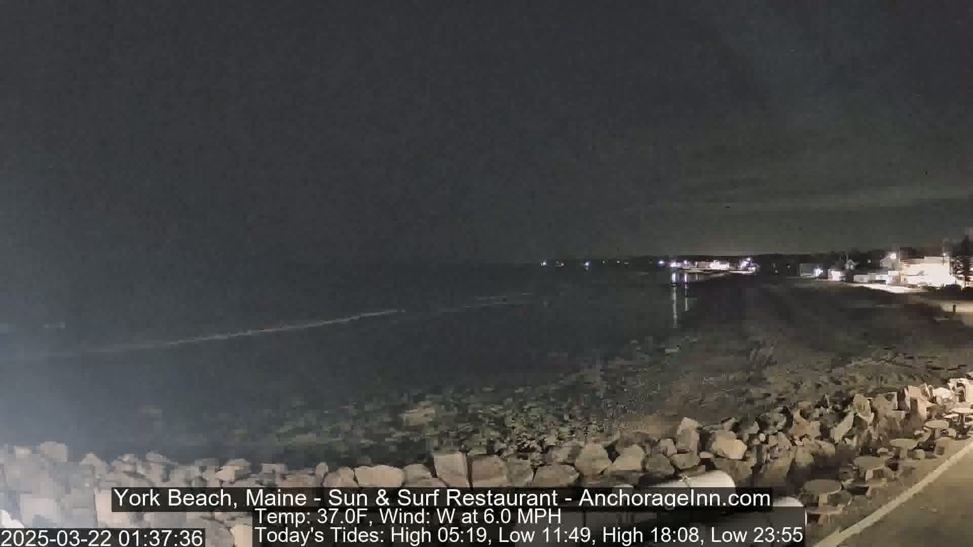 A dark, nighttime view of a rocky shoreline and calm ocean under a mostly clear sky.