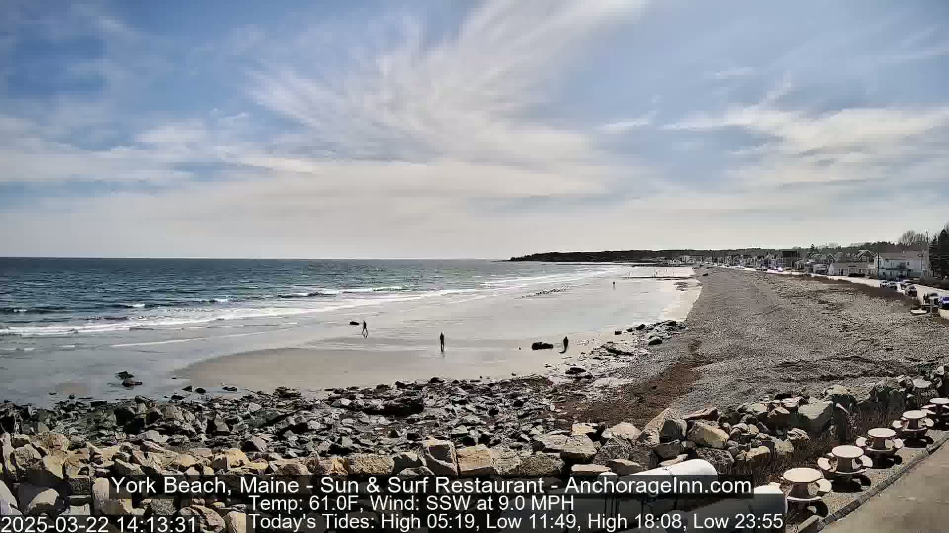 A long sandy beach stretches along a rocky coastline under a mostly sunny sky, with a few people walking along the shore and some buildings visible in the distance.