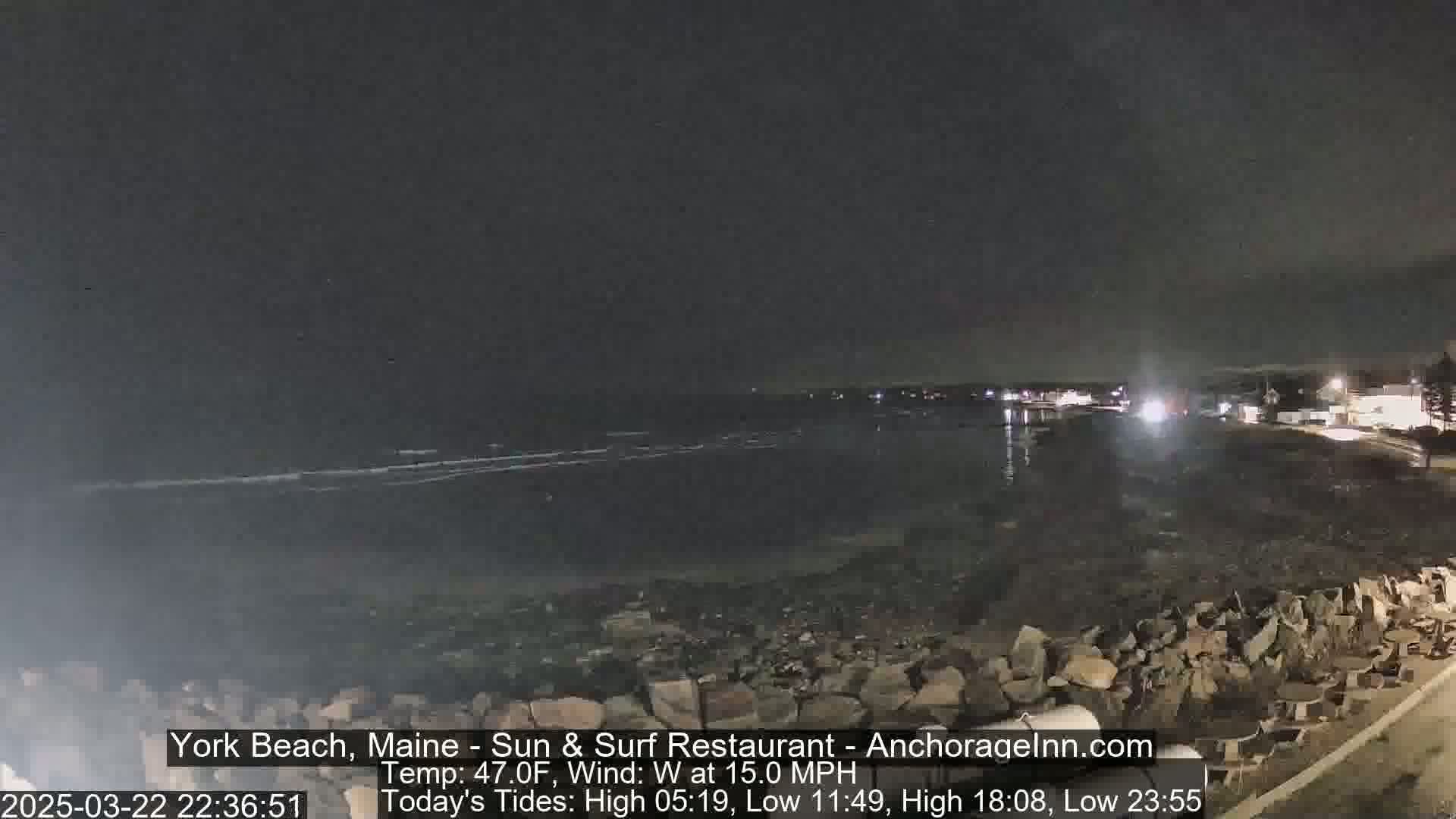 A nighttime coastal scene shows a dark ocean with small waves under a clear, dark sky, a rocky shoreline, and some distant lights.