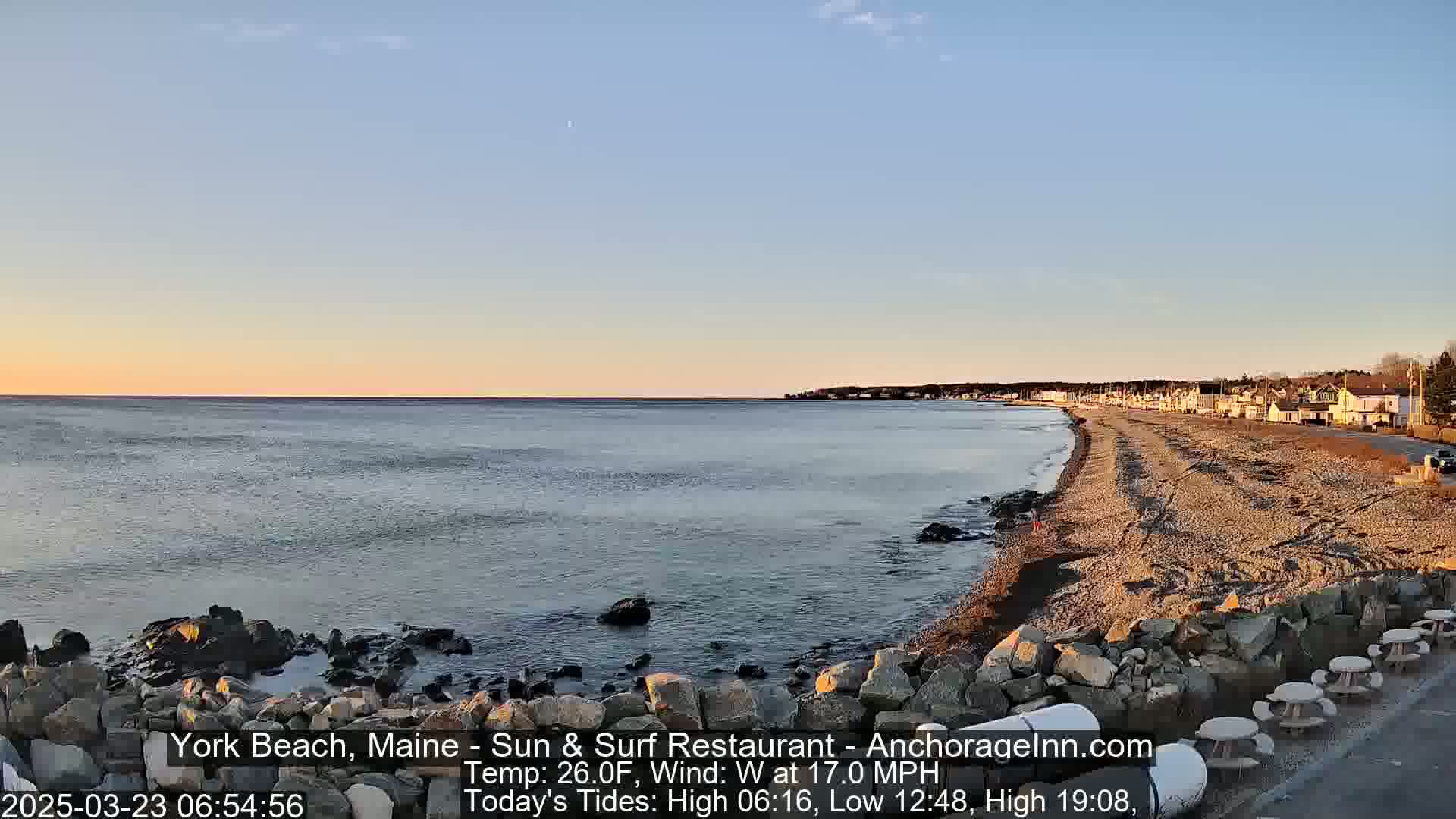 A calm ocean meets a sandy beach lined with houses under a clear, pale-blue sky at sunrise.