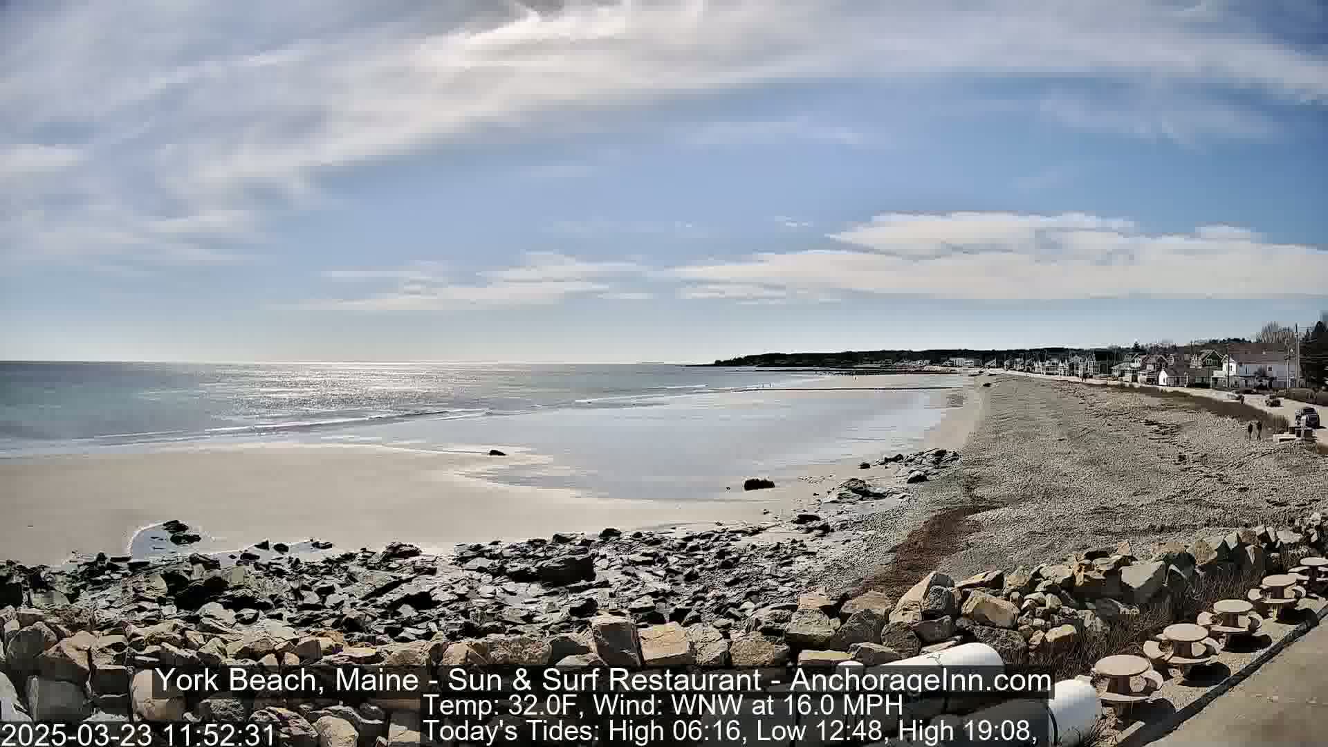 A sandy beach with rocky shoreline stretches towards a calm ocean under a partly cloudy sky.