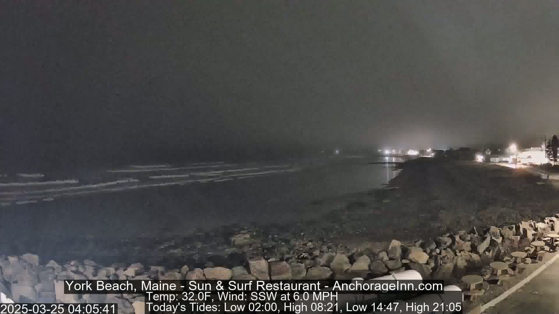 A nighttime, foggy view of a rocky shoreline with dark water and a calm ocean visible in the distance.