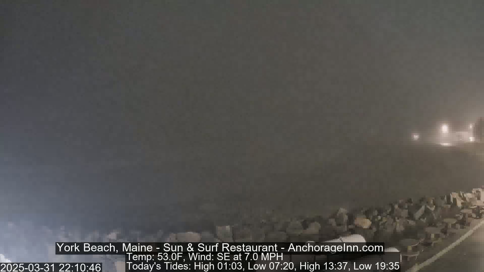 A foggy, nighttime coastal scene shows a rocky shoreline and dimly lit buildings in the distance.