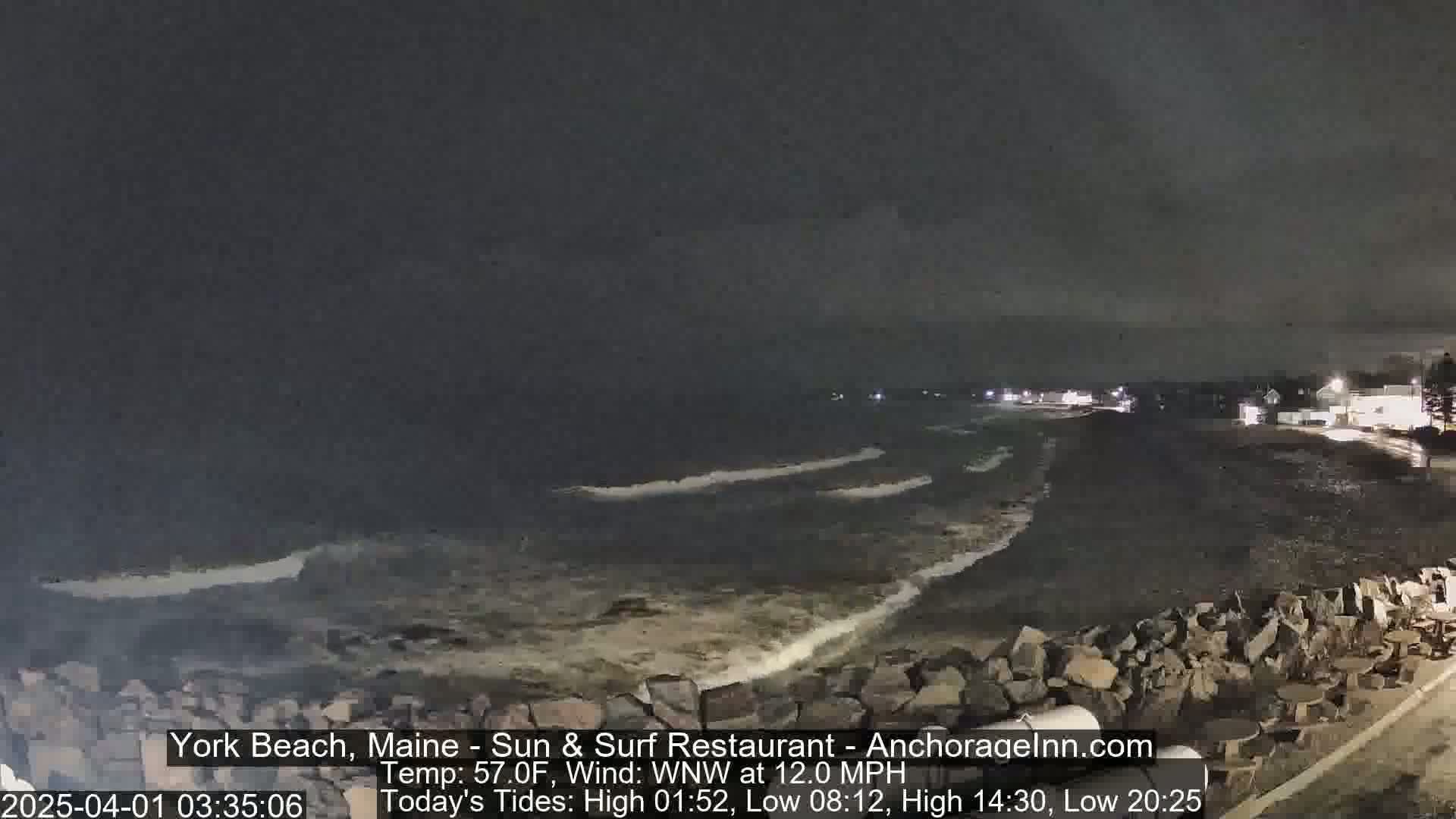 A nighttime view of a rocky beach with dark, wavy water under a cloudy sky.