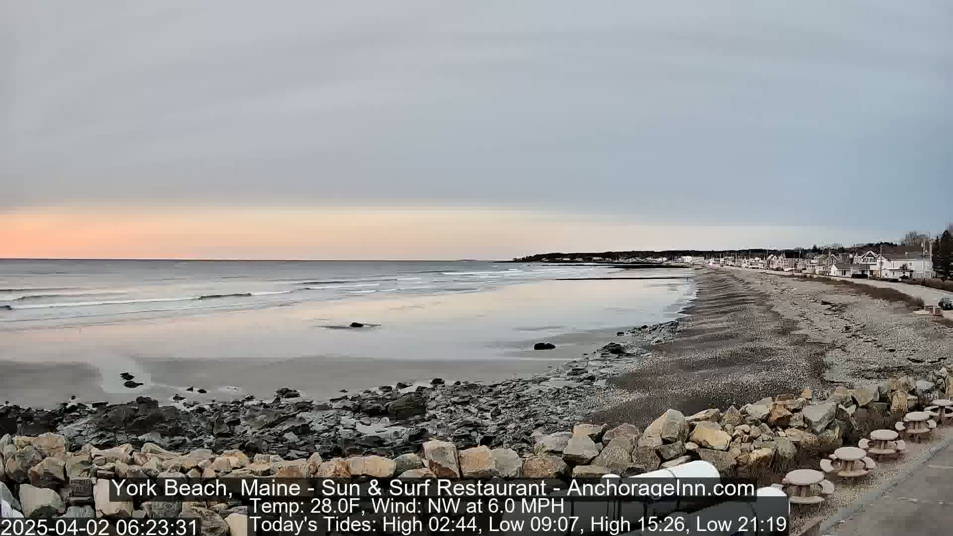 A rocky beach with calm waves under a pale sunrise sky shows a row of houses in the distance.