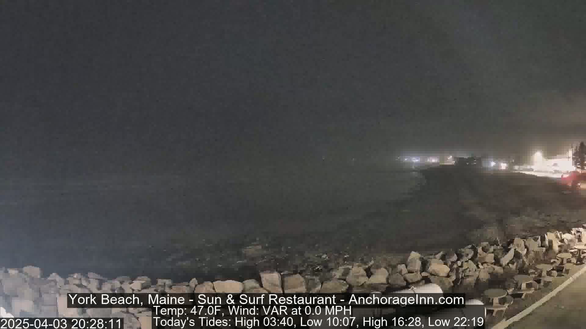 A nighttime, foggy coastal scene shows a dark beach with rocks and a roadway beyond.
