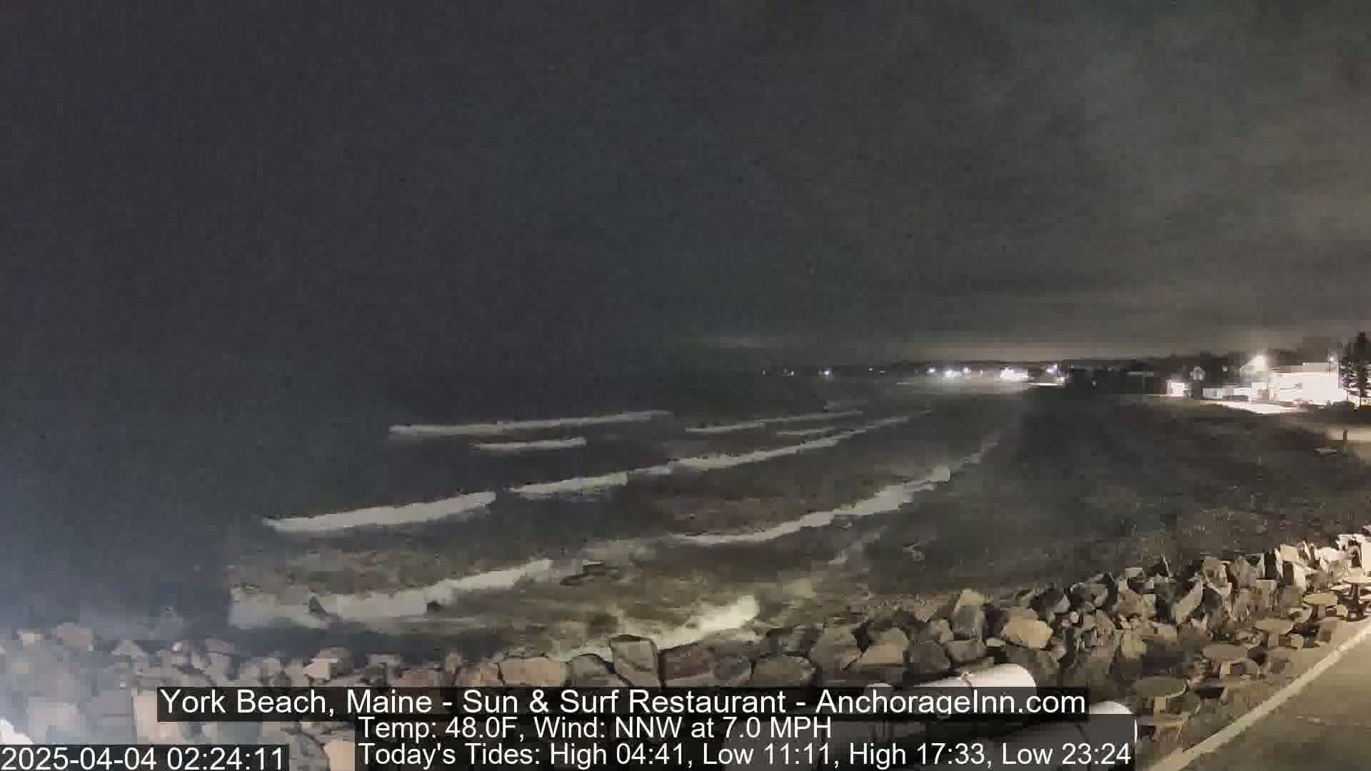 A nighttime beach scene shows dark, gentle waves lapping at a rocky shore under a mostly clear sky.