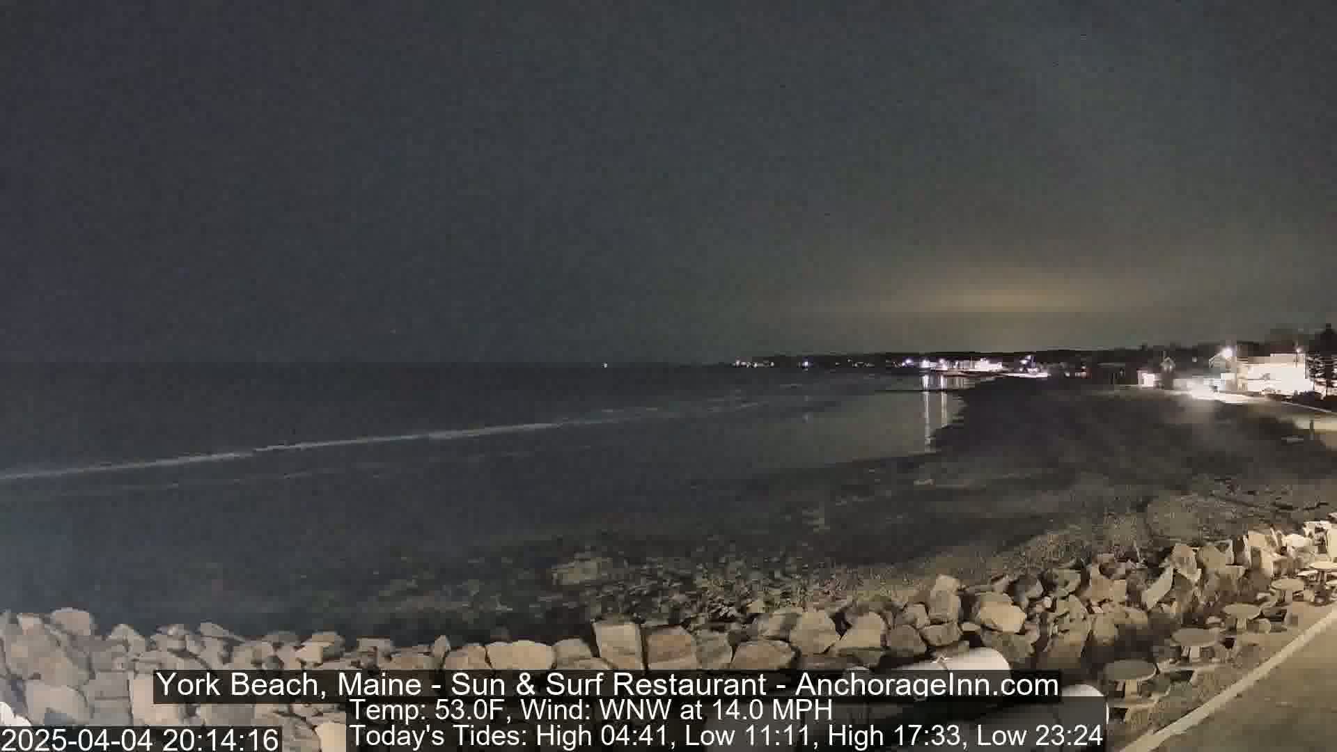 A dark, nighttime view of a rocky beach and calm ocean under a cloudy sky.