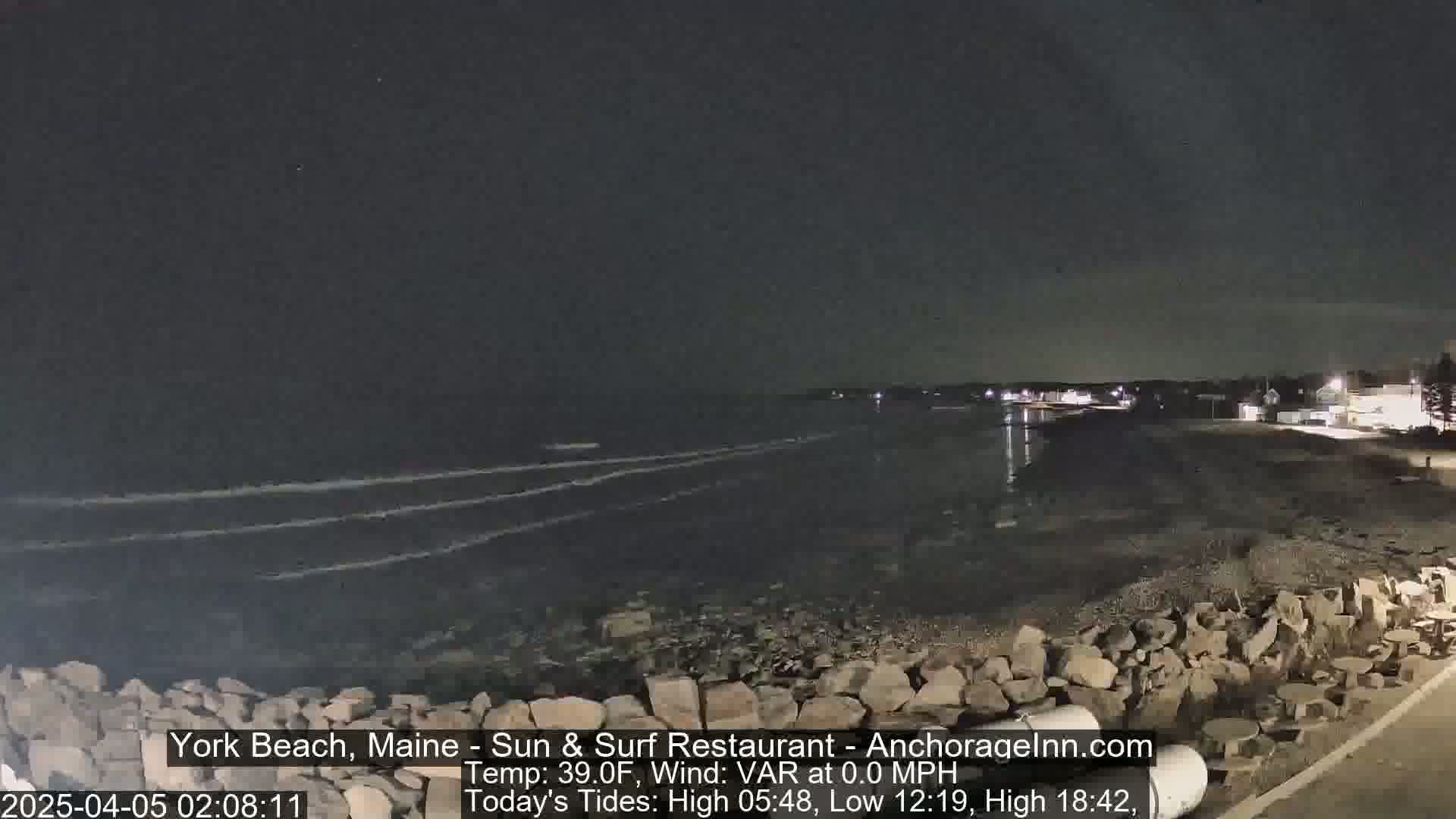 A dark, nighttime scene shows a rocky shoreline with calm ocean waves under a clear sky.