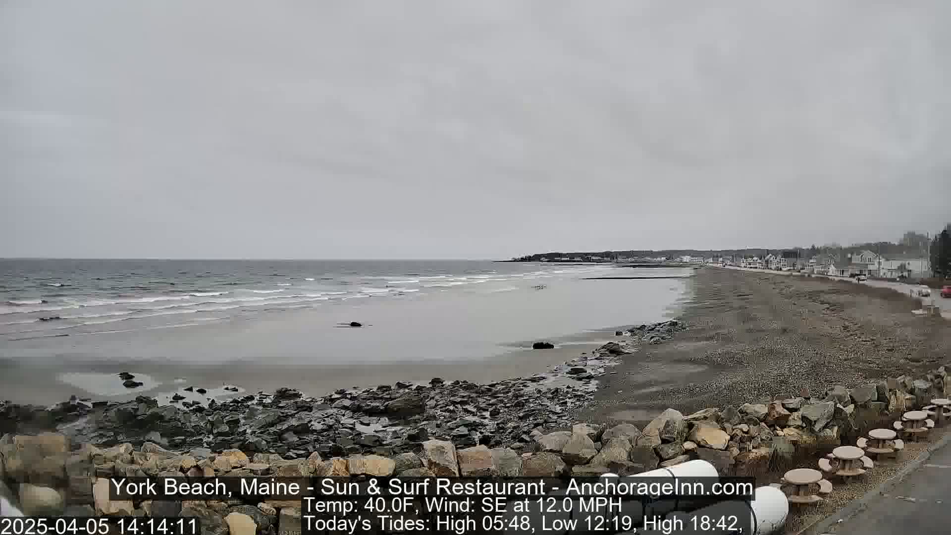 A gray, overcast day shows a beach with calm waves lapping the shore, a rocky shoreline, and a few buildings in the distance.