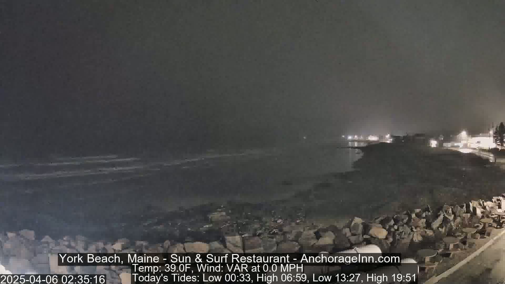 A dark, foggy nighttime scene shows a rocky shoreline with calm ocean waves and distant lights.