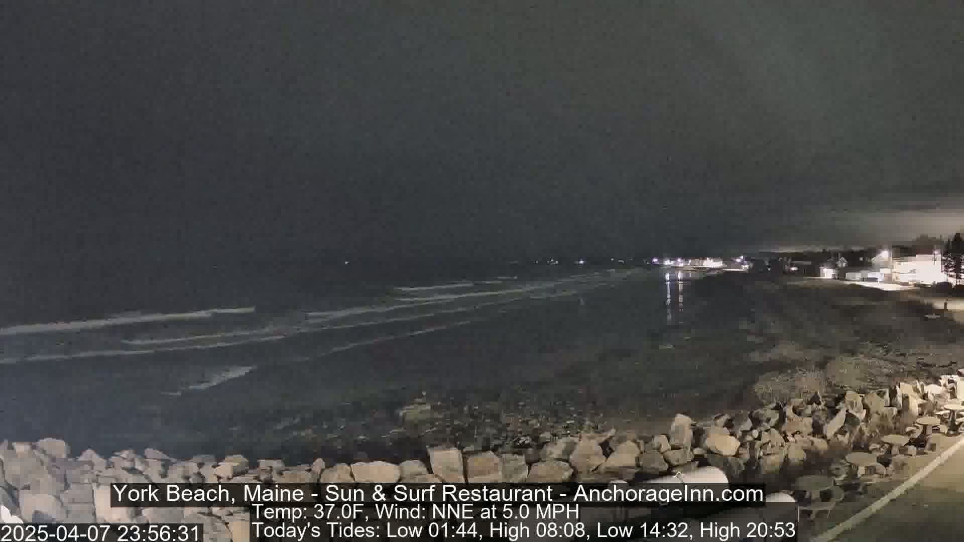 A nighttime view of a dark, rocky beach with a calm ocean under a cloudy sky.