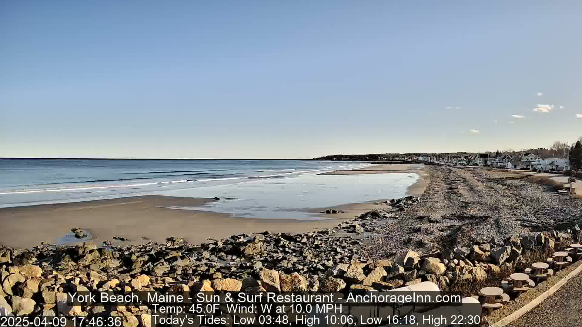 A sandy beach with small waves stretches along a rocky shoreline under a clear, sunny sky, with a line of buildings visible in the distance.