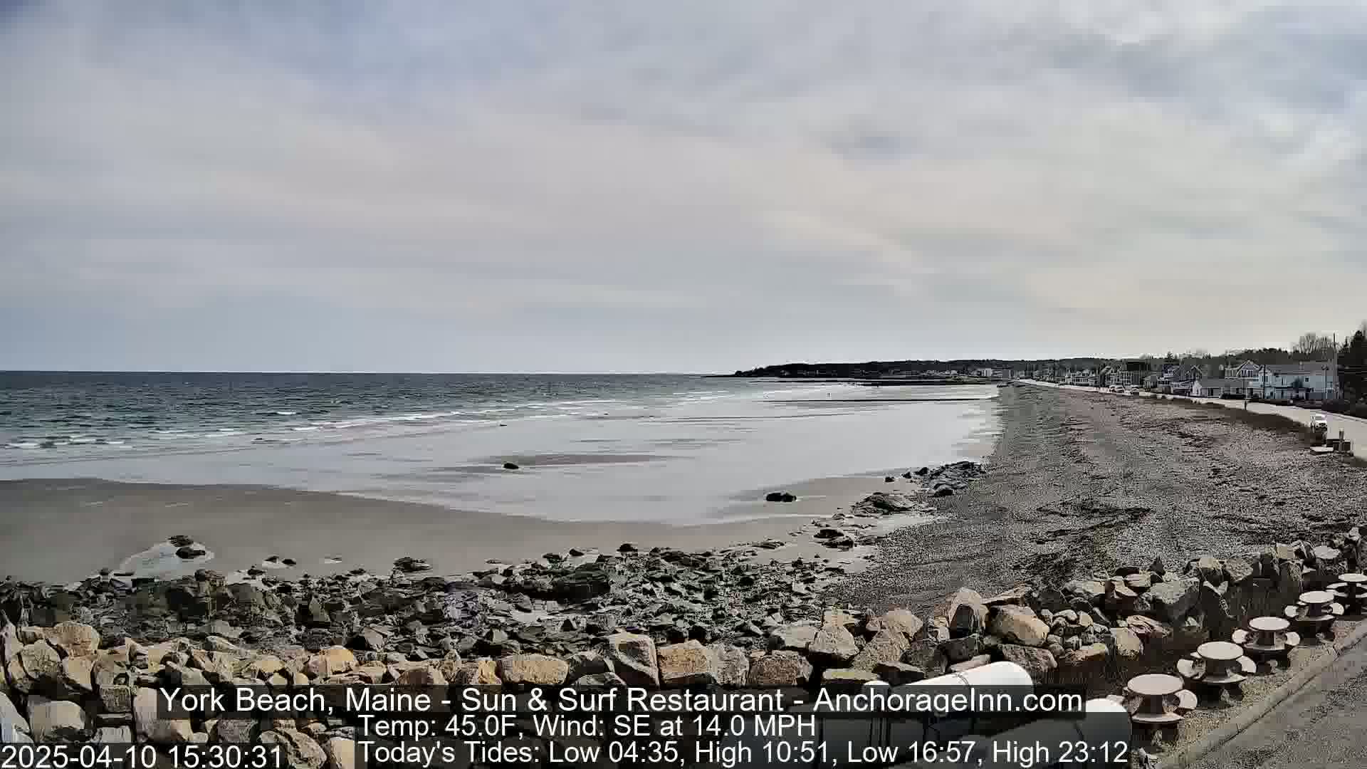 A rocky beach with a calm sea under a partly cloudy sky stretches to a shoreline of houses and trees.