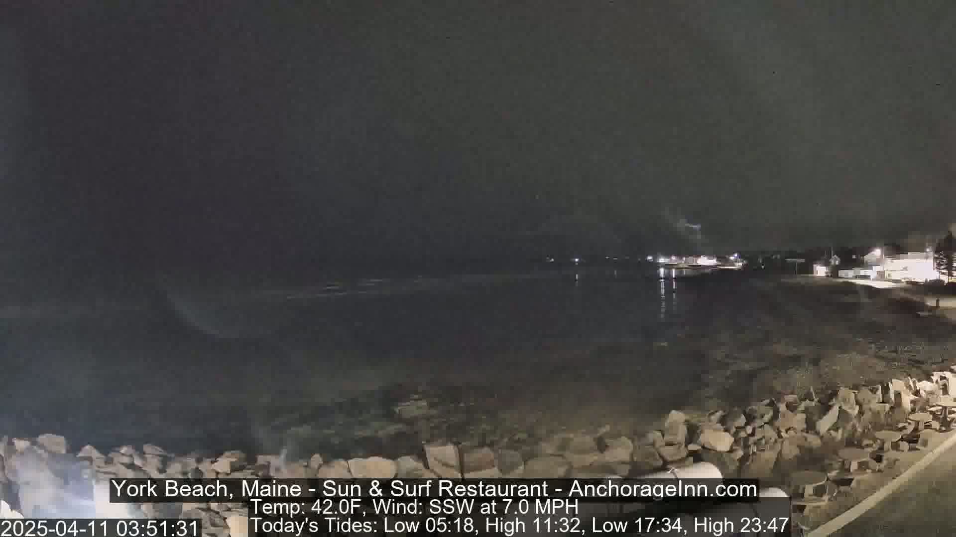 A dark nighttime view of a rocky beach and calm water, under a cloudy sky.