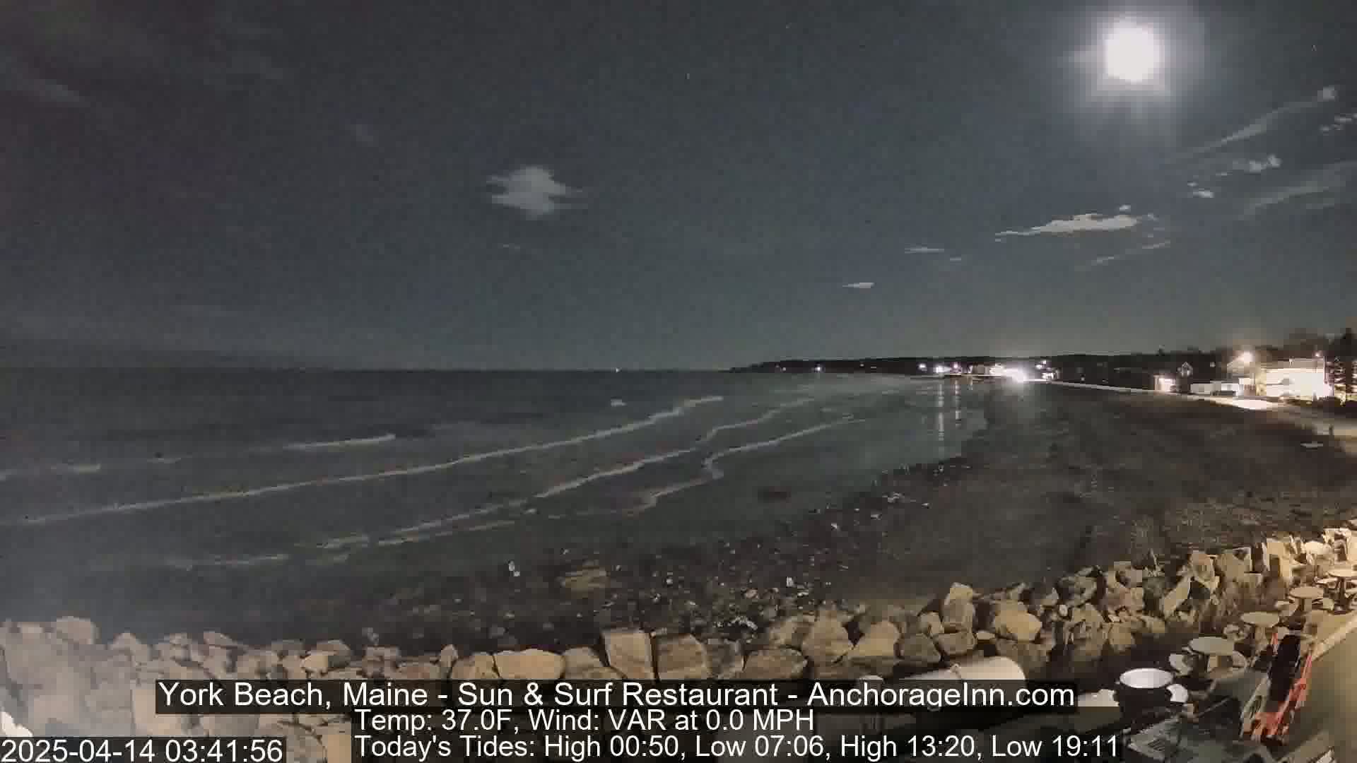 A nighttime beach scene under a bright moon shows a calm ocean with small waves lapping the shore, a dark sandy beach, and some rocks and outdoor furniture near the water's edge.