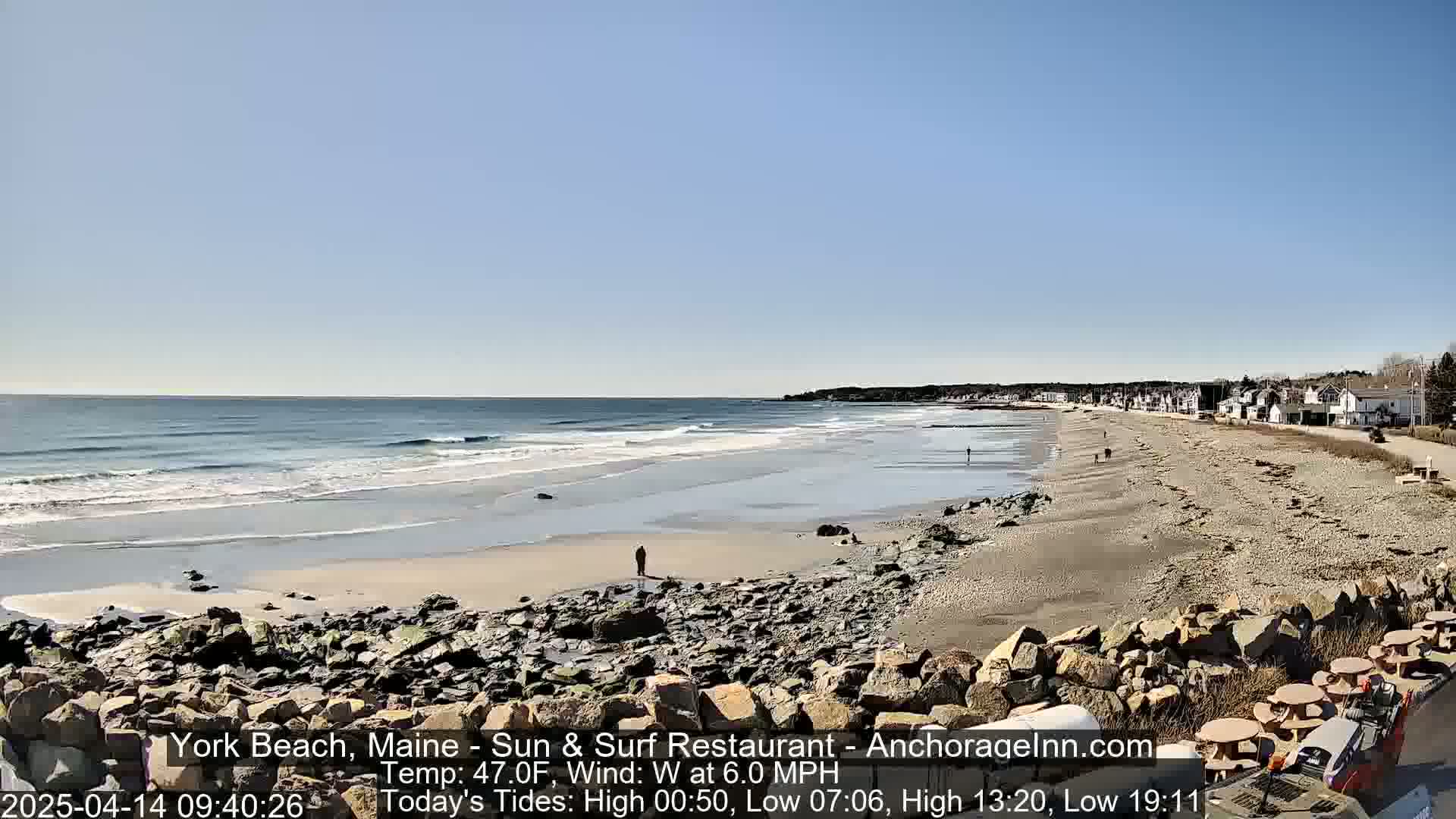A sandy beach with rocky shoreline stretches to a calm ocean under a clear blue sky.