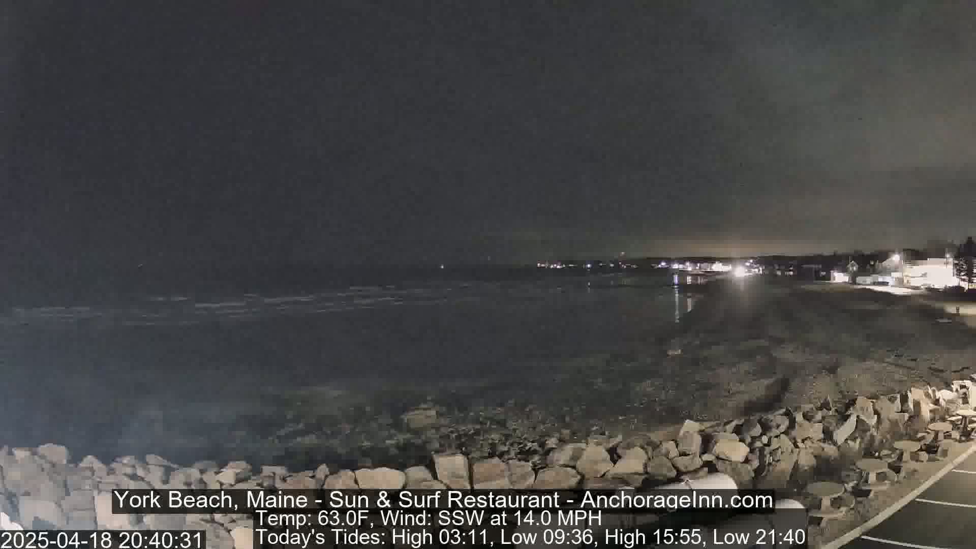 A nighttime beach scene with a rocky shore, dark water, and a town visible in the distance under a mostly clear, dark sky, with a strong wind.