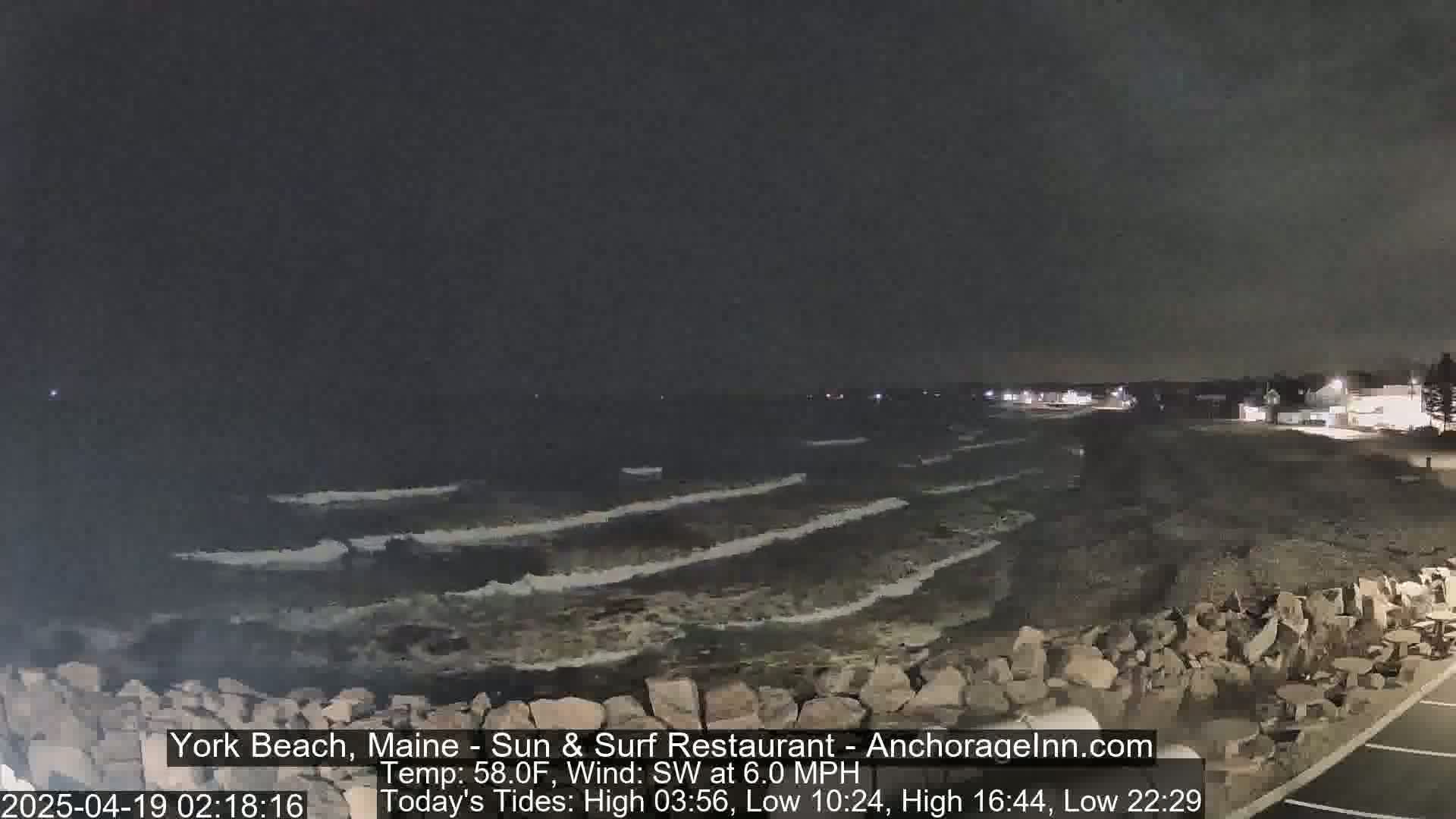 A dark, nighttime beach scene shows gently lapping waves on a rocky shore under a mostly clear sky.