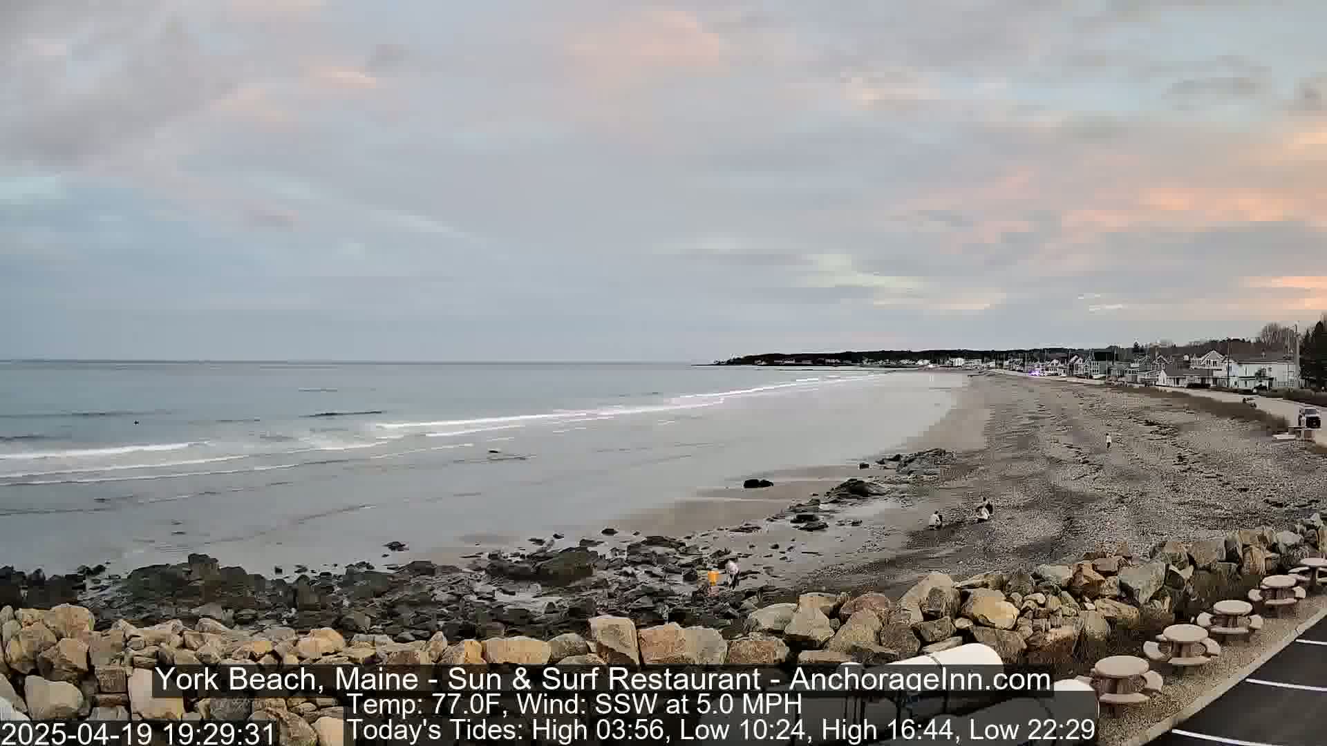 A long sandy beach with rocky outcrops stretches along a calm ocean under a mostly cloudy, pale sky.