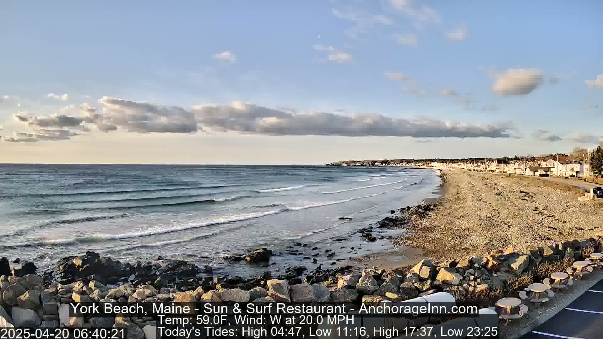 A sandy beach with small waves and a few houses in the distance under a partly cloudy sky.