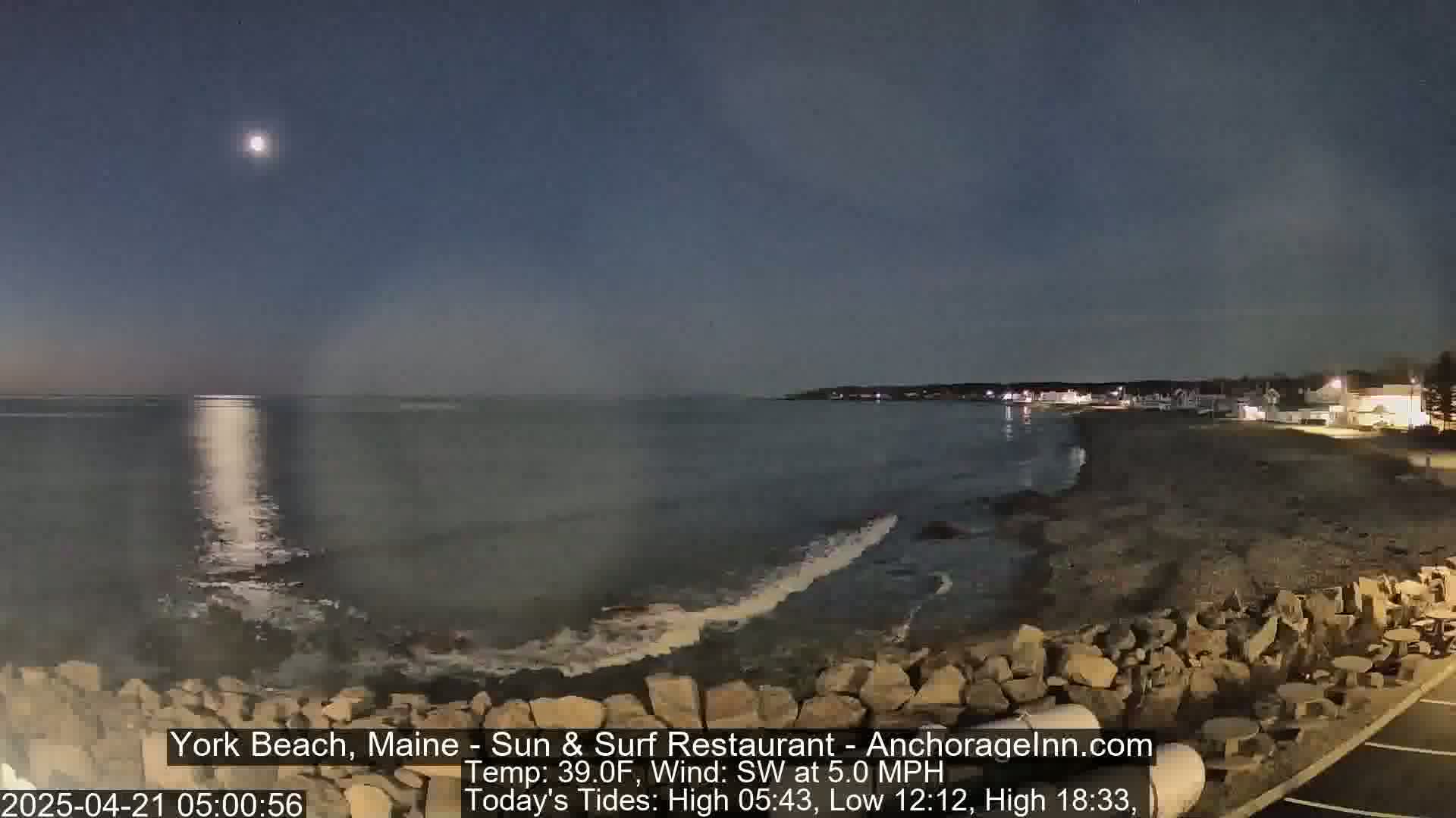 A moonlit ocean shore at night with gentle waves breaking on a rocky coastline, under a mostly clear sky.