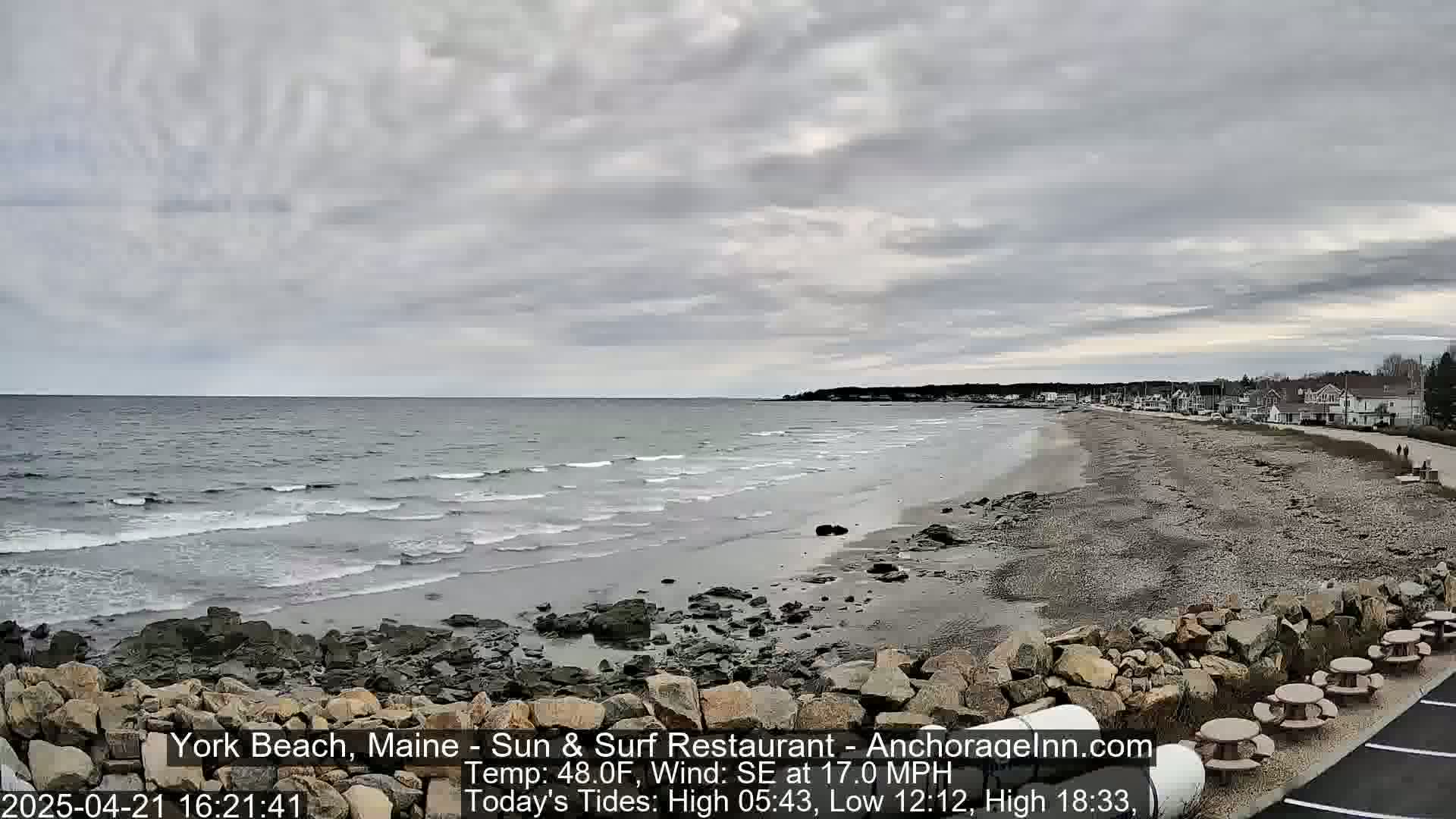 A wide shot of a beach with gentle waves under an overcast sky shows a rocky shoreline and buildings in the distance.
