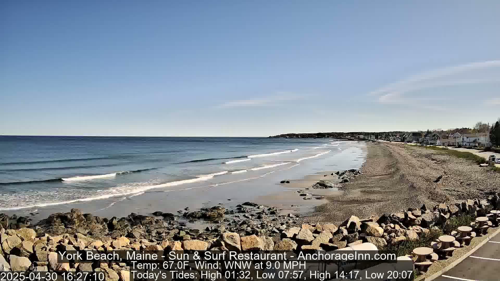 A sandy beach with small waves under a clear blue sky stretches to a line of buildings in the distance.