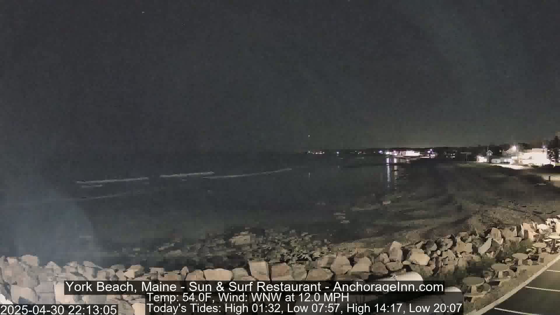 A nighttime coastal scene shows a dark body of water under a clear sky, with a rocky shoreline and distant lights.