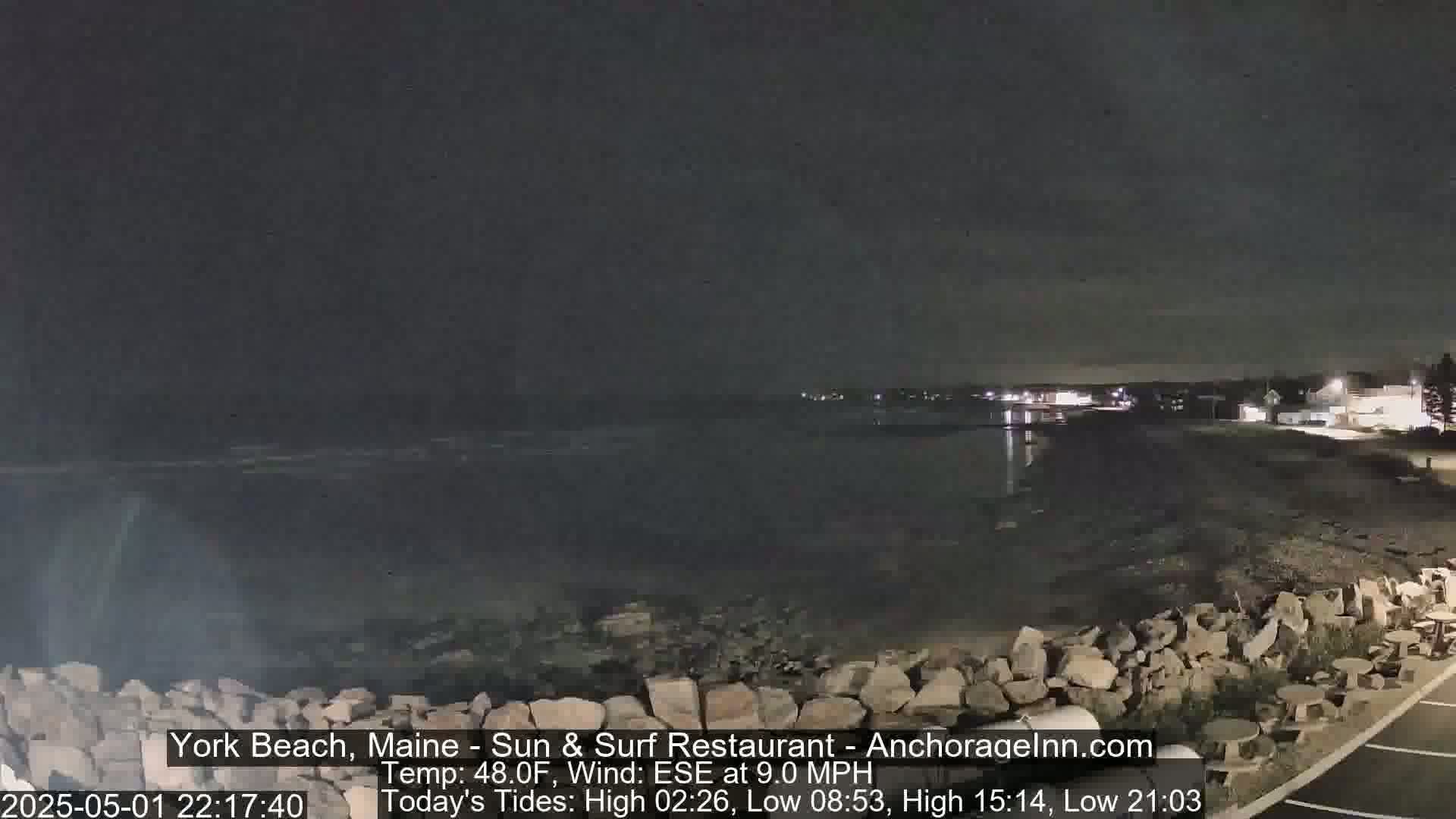 A nighttime, seaside view shows a dark body of water, a rocky shore, and a few distant lights.